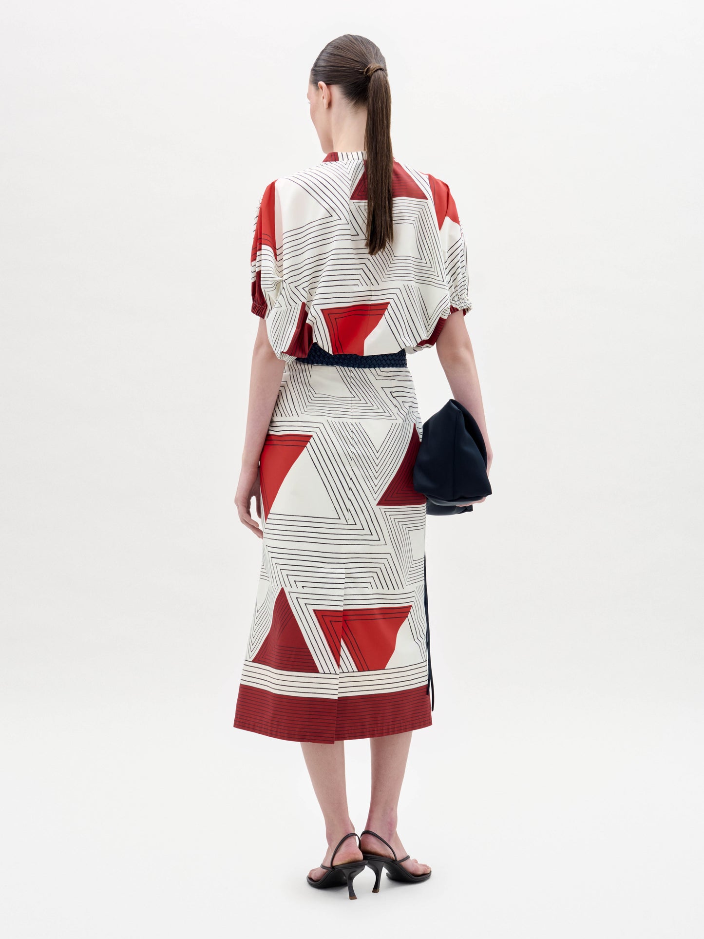 Una persona se coloca de espaldas con la falda Atira, una prenda de color crema con motivos geométricos rojos y líneas negras. Lleva sandalias de tacón negras, el pelo recogido en una coleta baja y un bolso de mano oscuro. El fondo es blanco.