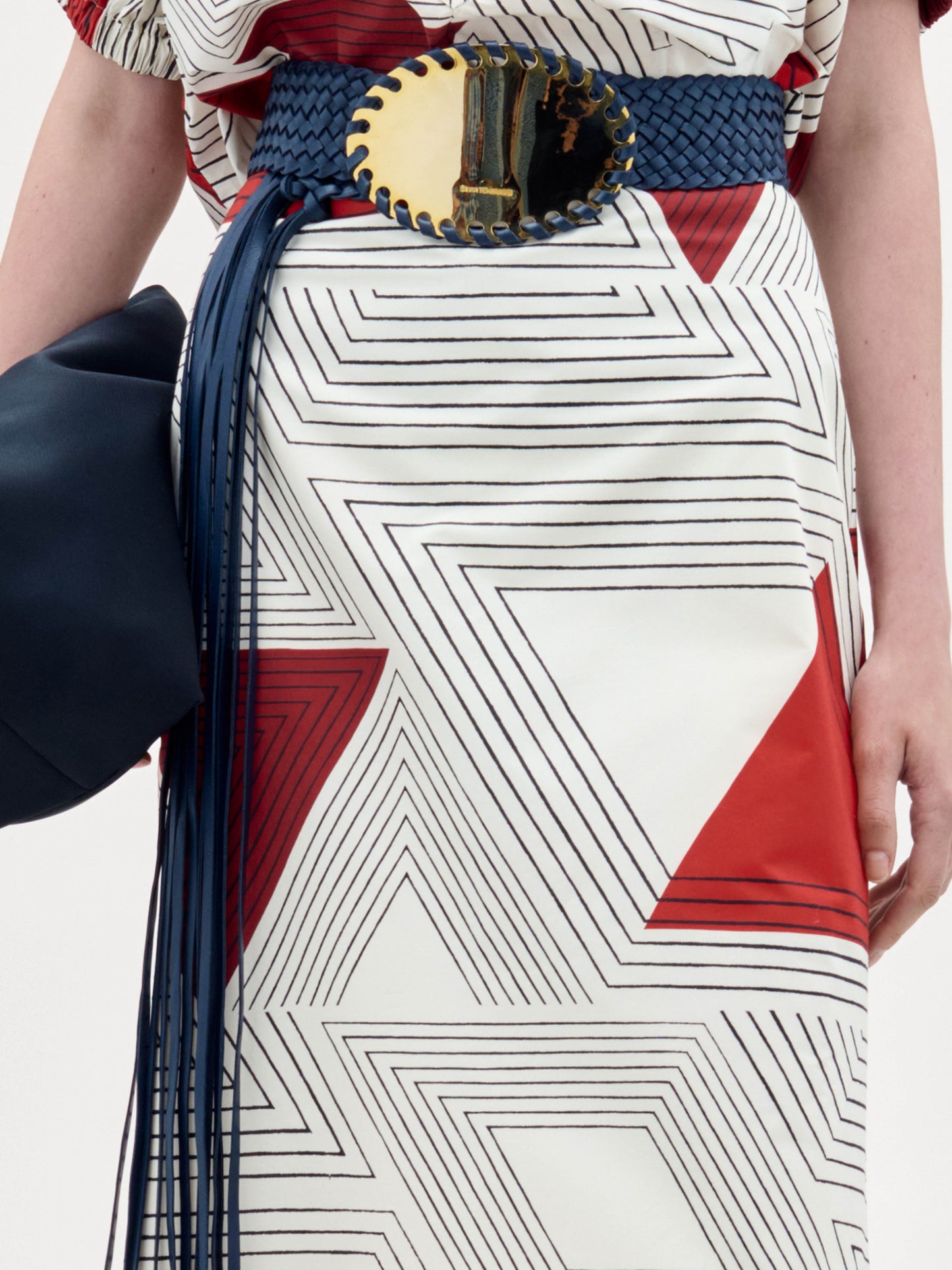Persona que lleva la falda Atira con estampado geométrico rojo y blanco, combinada con un cinturón ancho azul tejido con una gran hebilla ovalada dorada y un bolso azul oscuro.