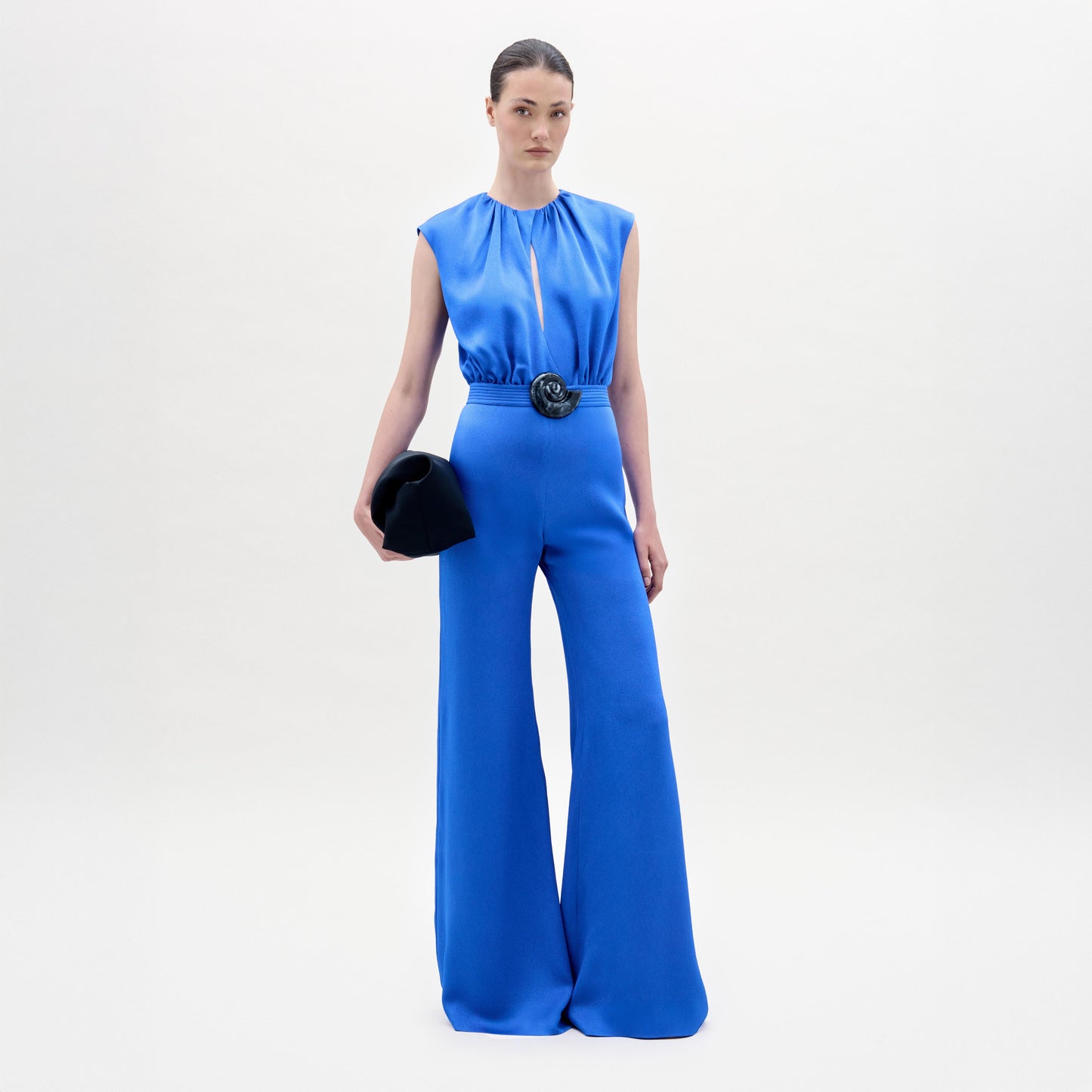 Una mujer posa de frente contra un fondo blanco, luciendo el Noel Jumpsuit en azul real, de bota ancha y con cinturón negro. Hecho en Colombia, combina el atuendo con clutch negro y lleva el cabello recogido elegantemente.
