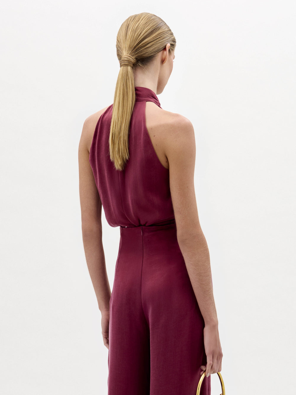 Una persona con el pelo rubio liso recogido en una coleta baja aparece de espaldas, llevando la blusa Shela sin mangas y de cuello alto en rojo intenso y sosteniendo un accesorio circular dorado sobre un fondo blanco liso.