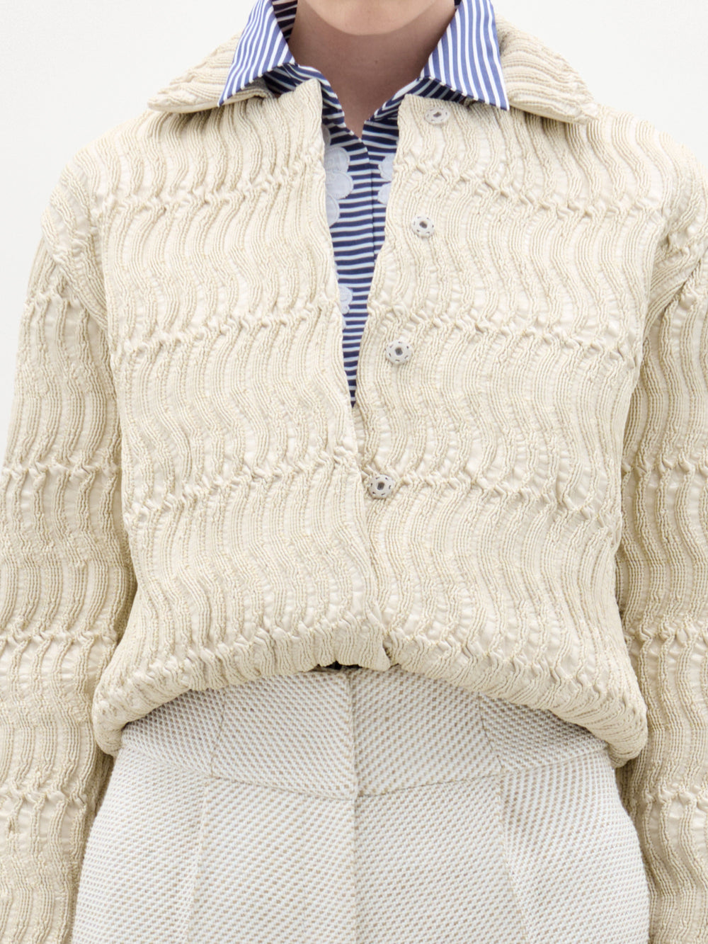 Una persona lleva la chaqueta Jaiden de color beige sobre una camisa de cuello a rayas azules y blancas y unos pantalones beige de cintura alta. La foto capta la parte superior del cuerpo desde los hombros hasta las caderas.