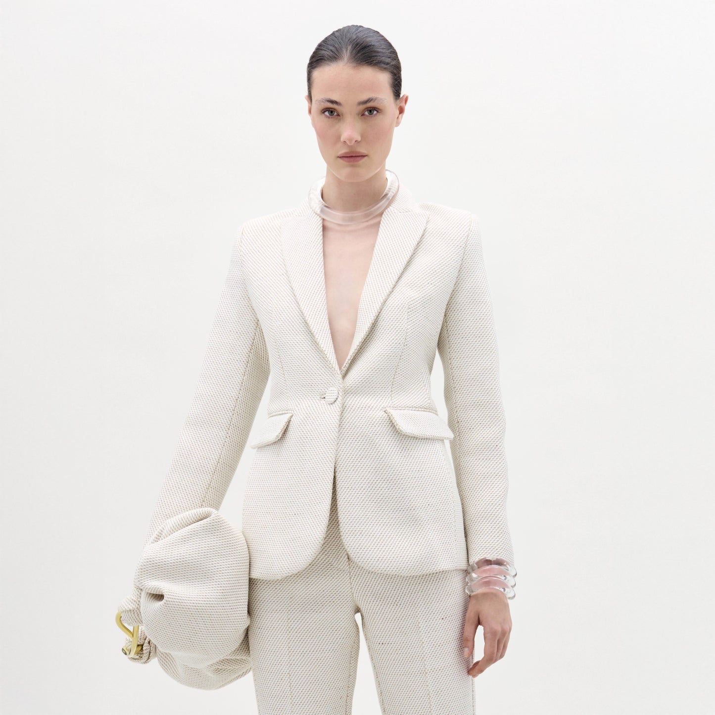 Una mujer posa sobre un fondo blanco con la chaqueta Rebeca, un blazer entallado de color hueso con textura y pantalones a juego. Lleva un bolso de mano grande de color hueso y brazaletes transparentes, resaltando una silueta elegante y estructurada.