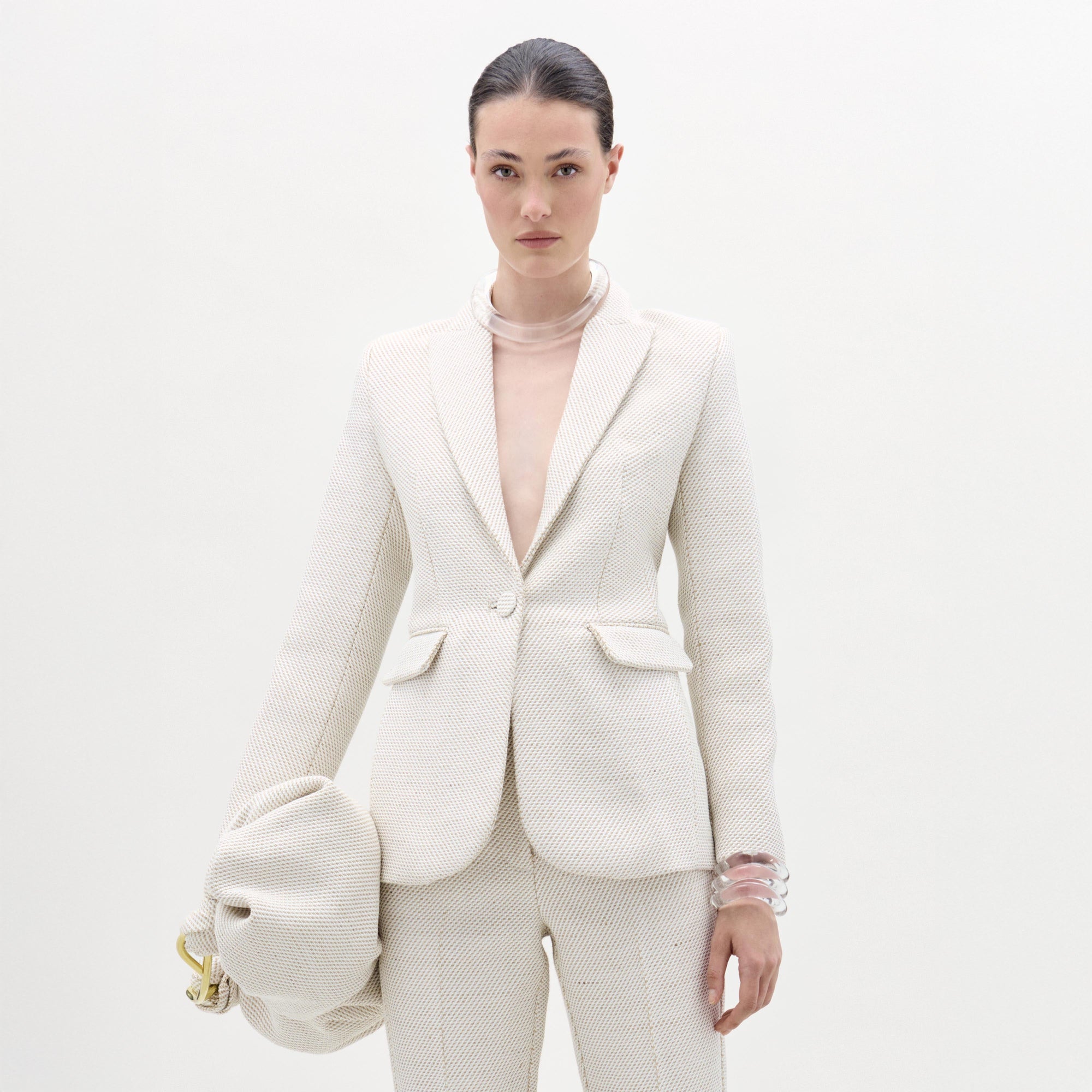 Una mujer posa sobre un fondo blanco con la chaqueta Rebeca, un blazer entallado de color hueso con textura y pantalones a juego. Lleva un bolso de mano grande de color hueso y brazaletes transparentes, resaltando una silueta elegante y estructurada.