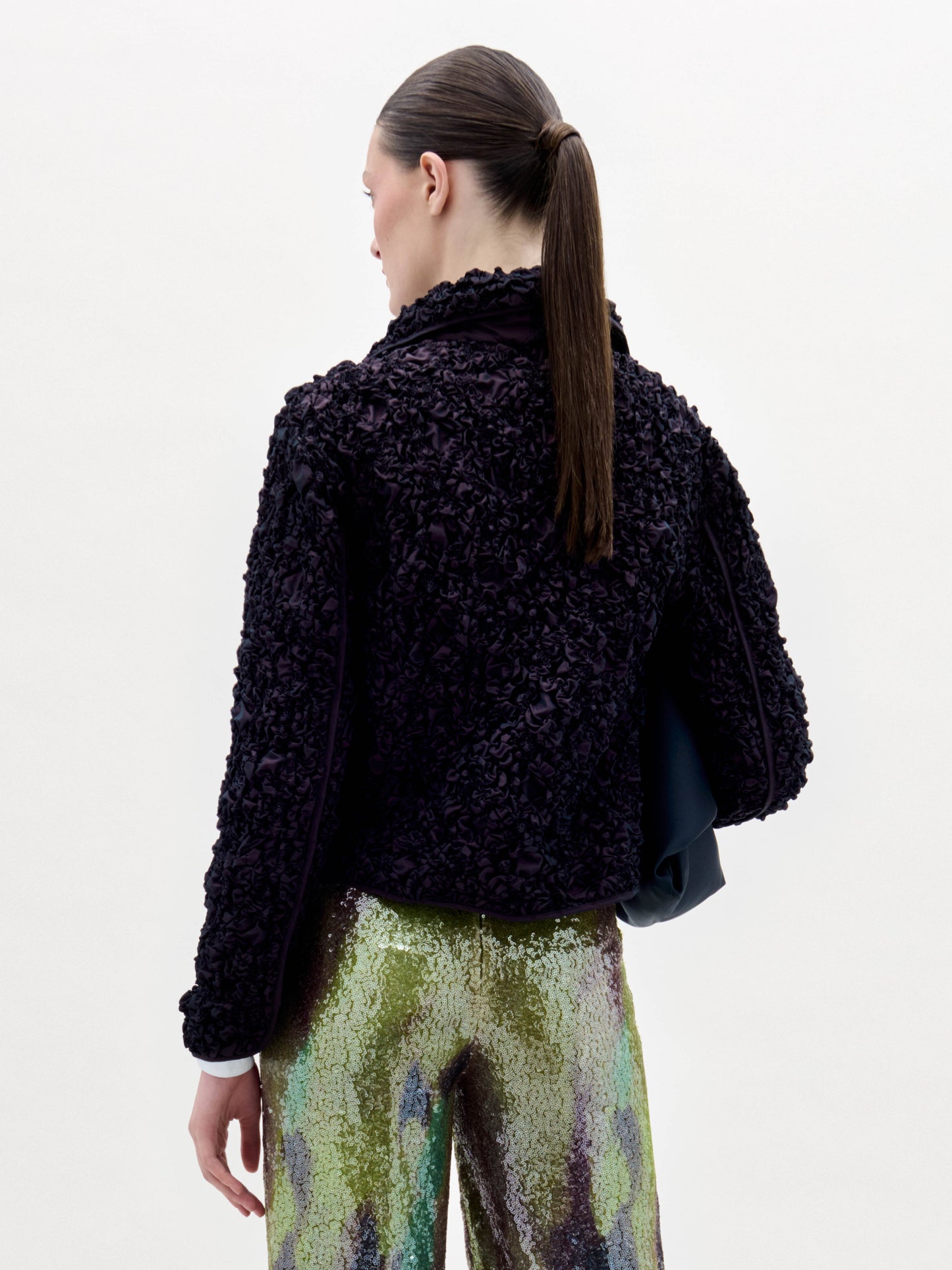 Una persona se encuentra de espaldas con la chaqueta Jamari -una prenda negra texturizada- combinada con unos pantalones brillantes, verdes y morados iridiscentes. Lleva el pelo oscuro recogido sobre un fondo blanco liso.