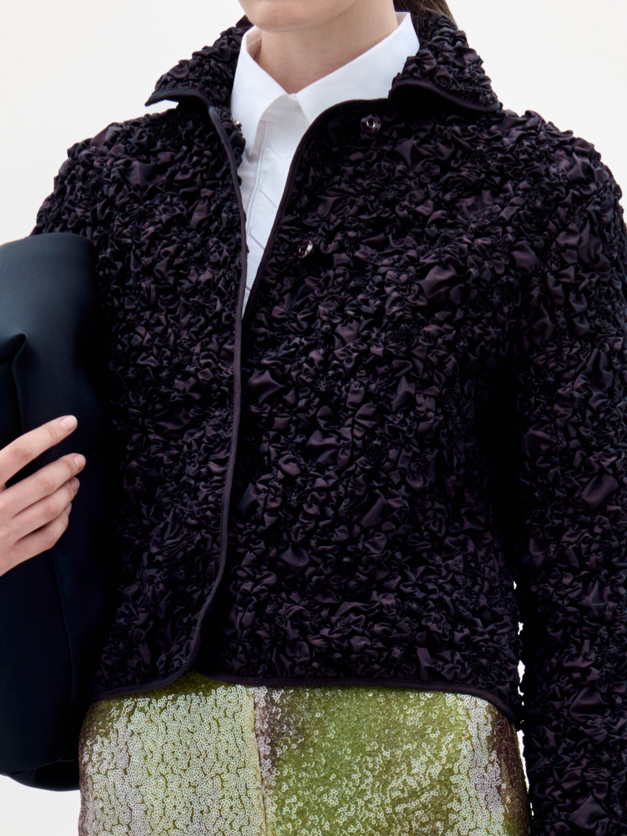 Una persona viste la chaqueta Jamari -una chaqueta negra con textura- sobre una camisa blanca de cuello, lleva un bolso de mano oscuro y viste una falda de lentejuelas verdes y plateadas, en la que sólo se ve la parte superior del cuerpo.