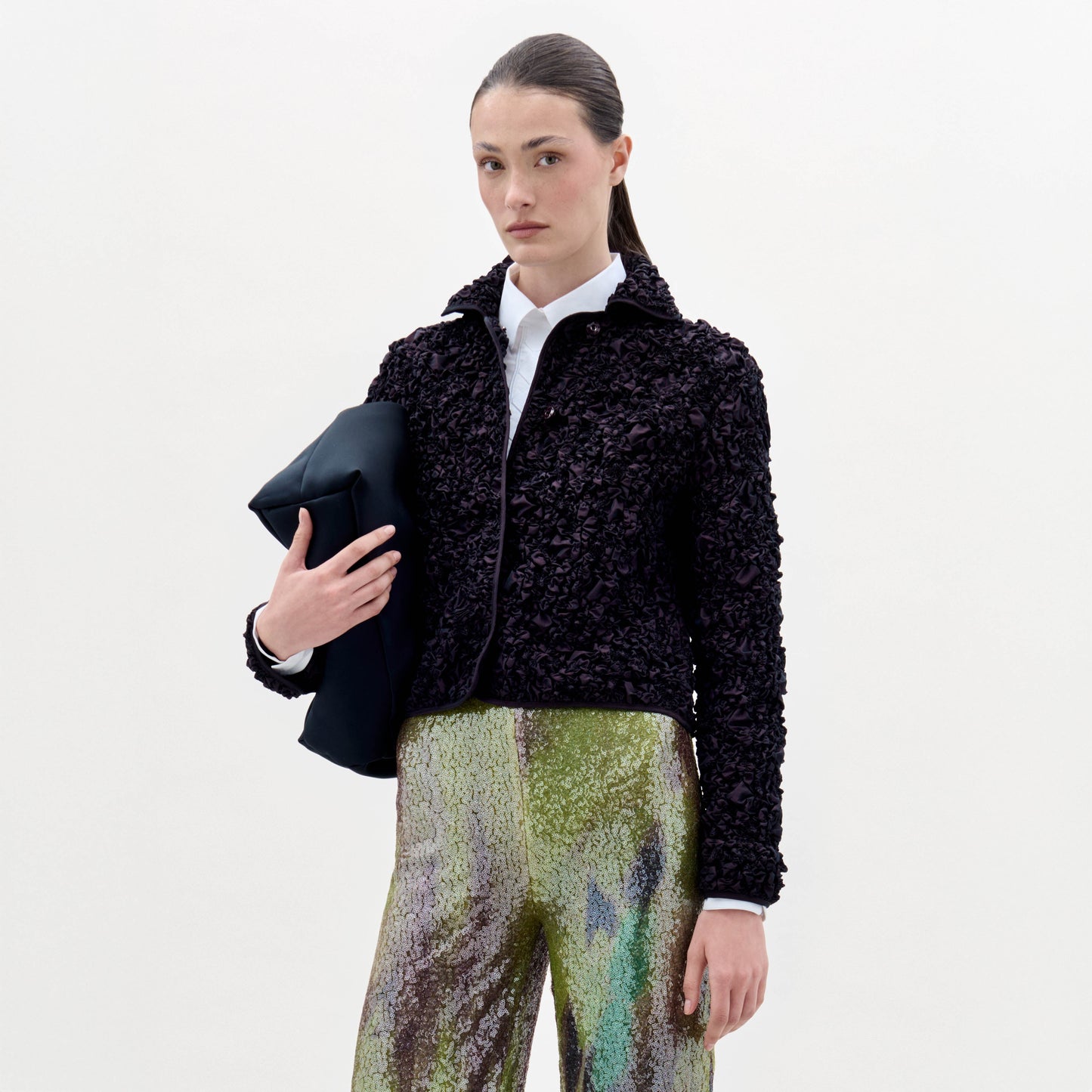 Una mujer con el pelo castaño liso echado hacia atrás lleva la chaqueta Jamari sobre una camisa blanca y unos pantalones con estampado verde brillante, y sostiene una prenda oscura doblada, de pie sobre un fondo blanco liso.
