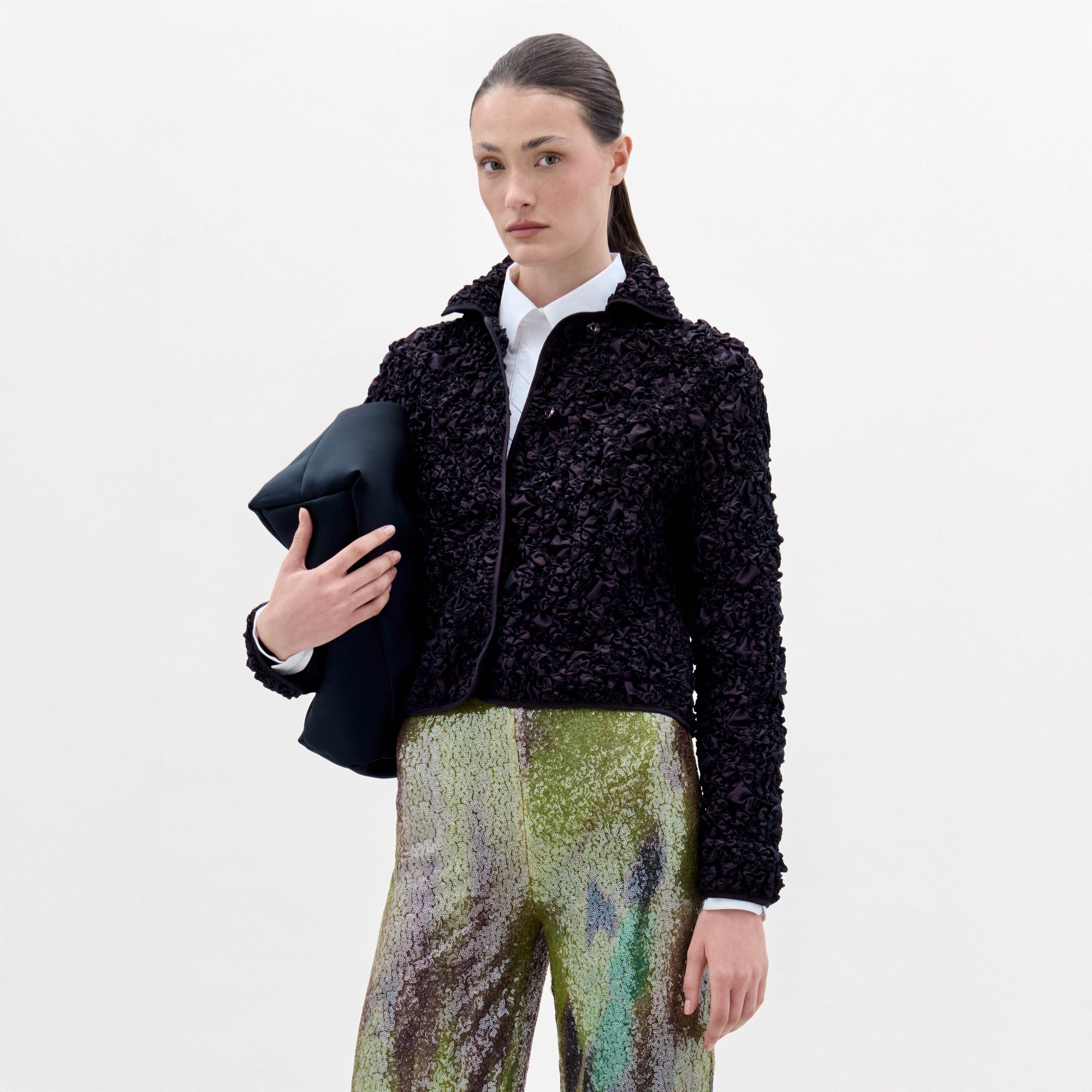 Una mujer con el pelo castaño liso echado hacia atrás lleva la chaqueta Jamari sobre una camisa blanca y unos pantalones con estampado verde brillante, y sostiene una prenda oscura doblada, de pie sobre un fondo blanco liso.