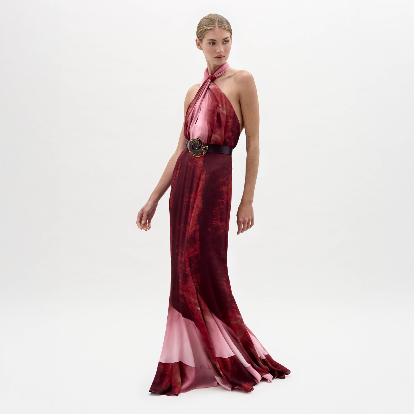 Una mujer posa sobre un fondo blanco con el vestido Raisa, sin mangas y largo hasta el suelo, de color rojo y rosa, con escote halter y cinturón negro con hebilla decorativa.