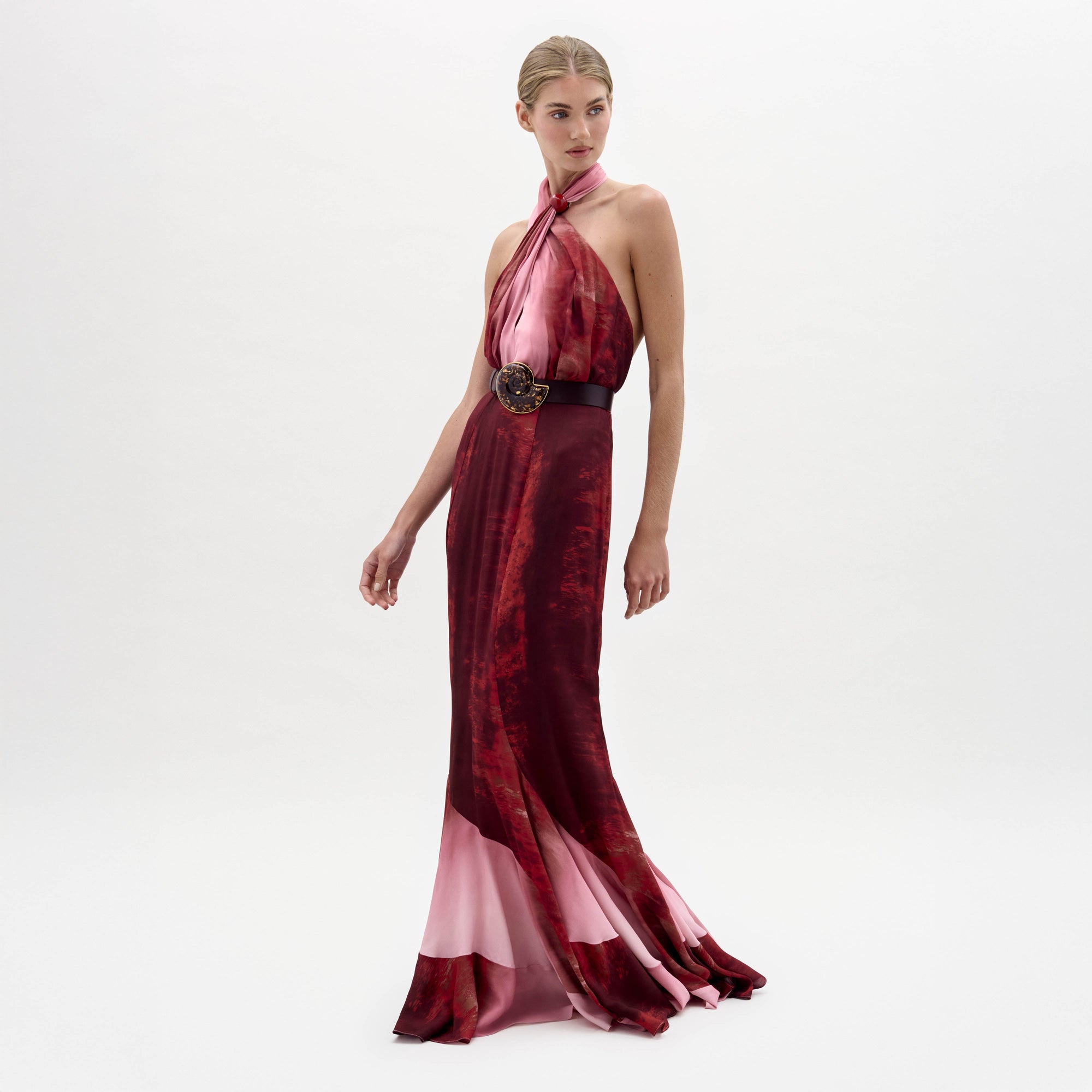 Una mujer posa sobre un fondo blanco con el vestido Raisa, sin mangas y largo hasta el suelo, de color rojo y rosa, con escote halter y cinturón negro con hebilla decorativa.