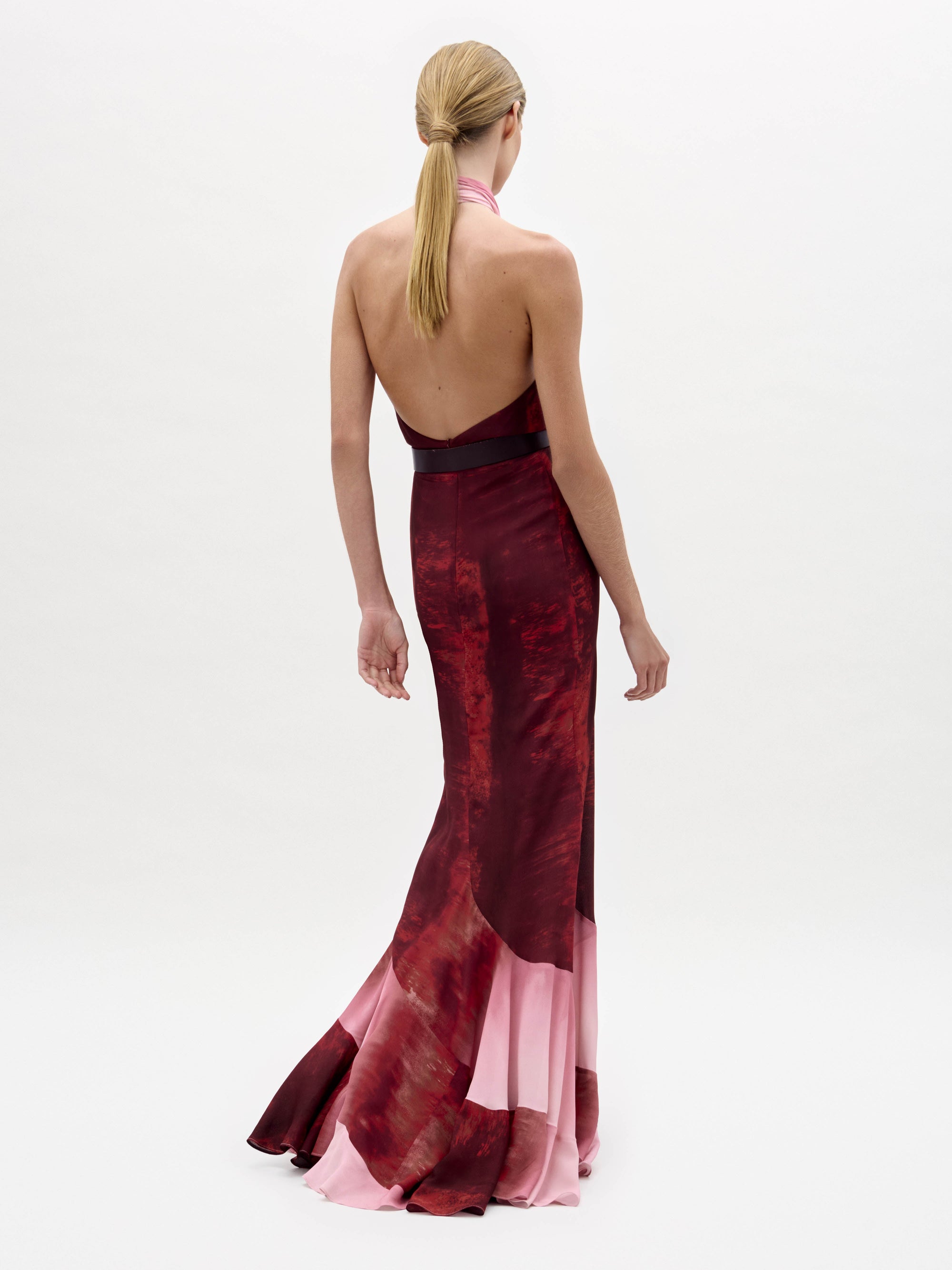 Una persona con el pelo rubio liso recogido en una coleta baja posa con el vestido Raisa, un modelo sin espalda, largo hasta el suelo y de corte halter en tonos rojos y rosas, sobre un fondo blanco liso.