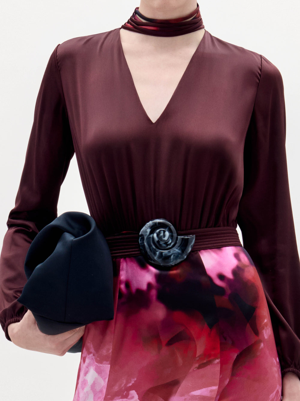 Una persona lleva el Jailyn Dress en burdeos oscuro con una gargantilla a juego, cinturón floral negro, falda en degradado rosa-rojo y sostiene un clutch negro doblado. No se ve la cara.