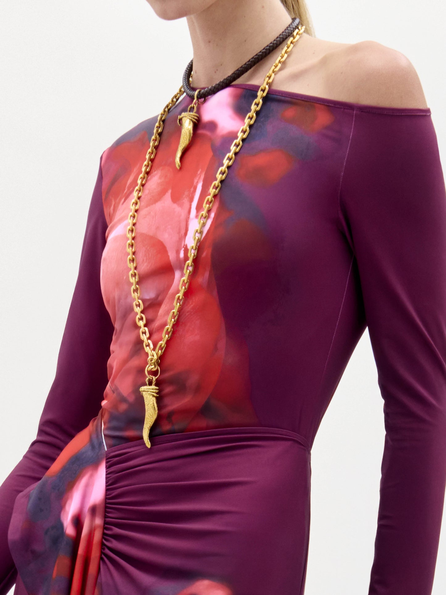 Una persona lleva la blusa Sabra, un top morado y rojo de manga larga y hombros descubiertos con frunces en la cintura. Lleva dos collares de cadena dorada con colgantes en forma de cuerno y el pelo recogido.