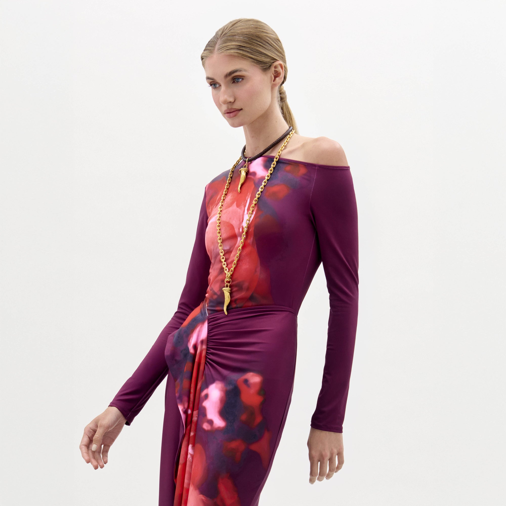 Una mujer con el pelo rubio peinado con elegancia lleva la blusa Sabra, un top de manga larga y hombros descubiertos con un estampado abstracto morado y rojo. La combina con un collar largo dorado sobre un fondo blanco liso.