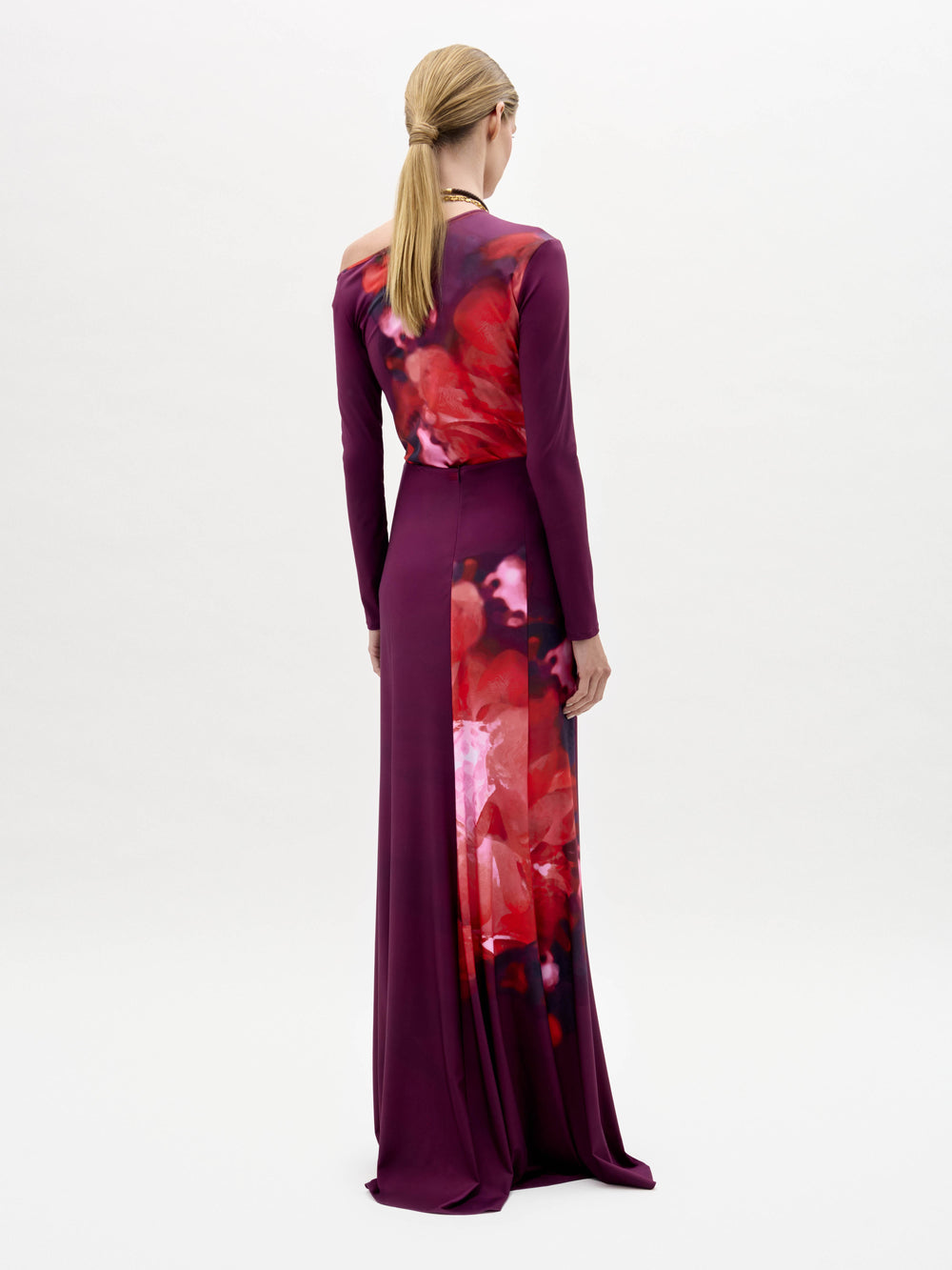 Una persona con el pelo rubio recogido en una coleta baja lleva la falda Meissa, una falda larga de color magenta con un estampado floral abstracto rojo y rosa en la espalda, sobre un fondo blanco liso.