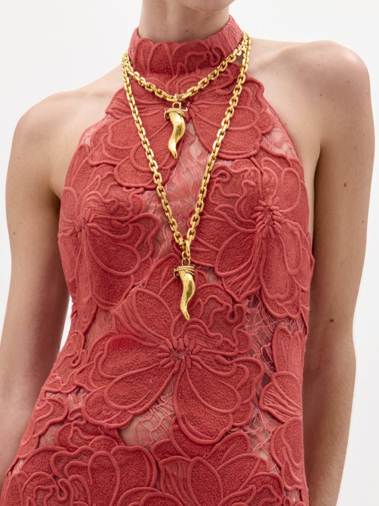 Una persona lleva el vestido Mab -un vestido de encaje coral sin mangas y cuello alto con llamativos motivos florales- combinado con dos collares de cadena de oro, cada uno con un colgante de guindilla dorada.