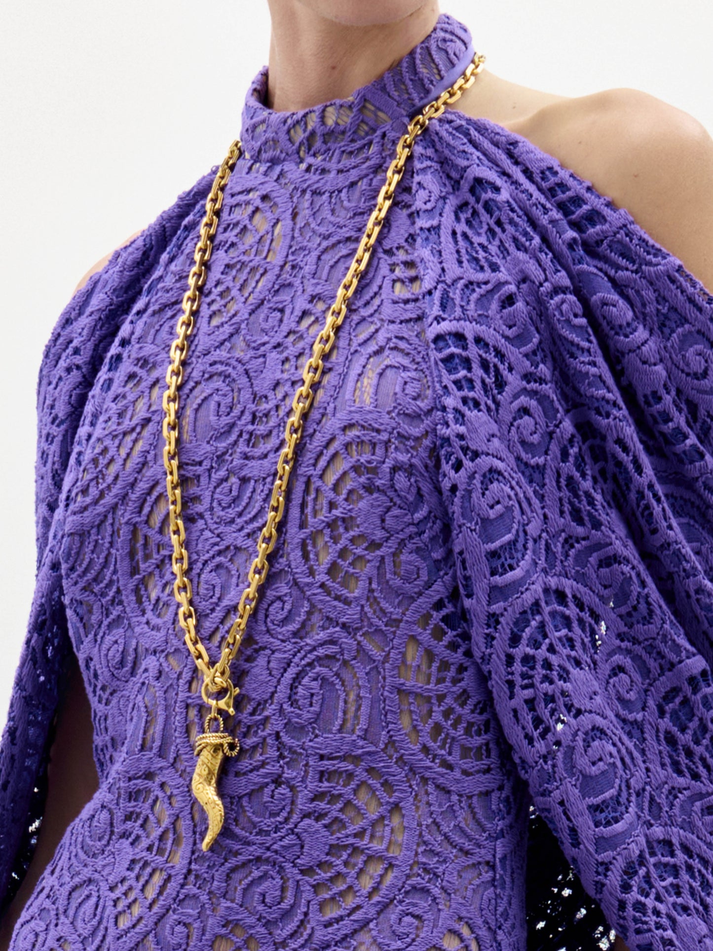Una persona con un top de encaje morado de cuello alto y hombros descubiertos, adornado con el Martina Long Necklace, una cadena de oro con un colgante dorado en forma de cuerno.