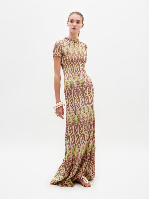 A person standing and posing in the Yara Dress Zigzag Neon, a floor-length dress with a vibrant zigzag design, adorned with large bracelet accessories and wearing flat sandals, against a white background. Pre-order now to ensure your Yara Dress Zigzag Neon ships by February 15th.