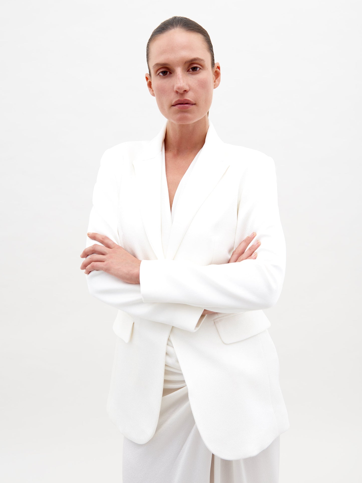 A person with a neutral expression is wearing the Rebeca Jacket White and white pants, standing against a plain background with arms crossed, available for pre-order starting February 15th, 2025.