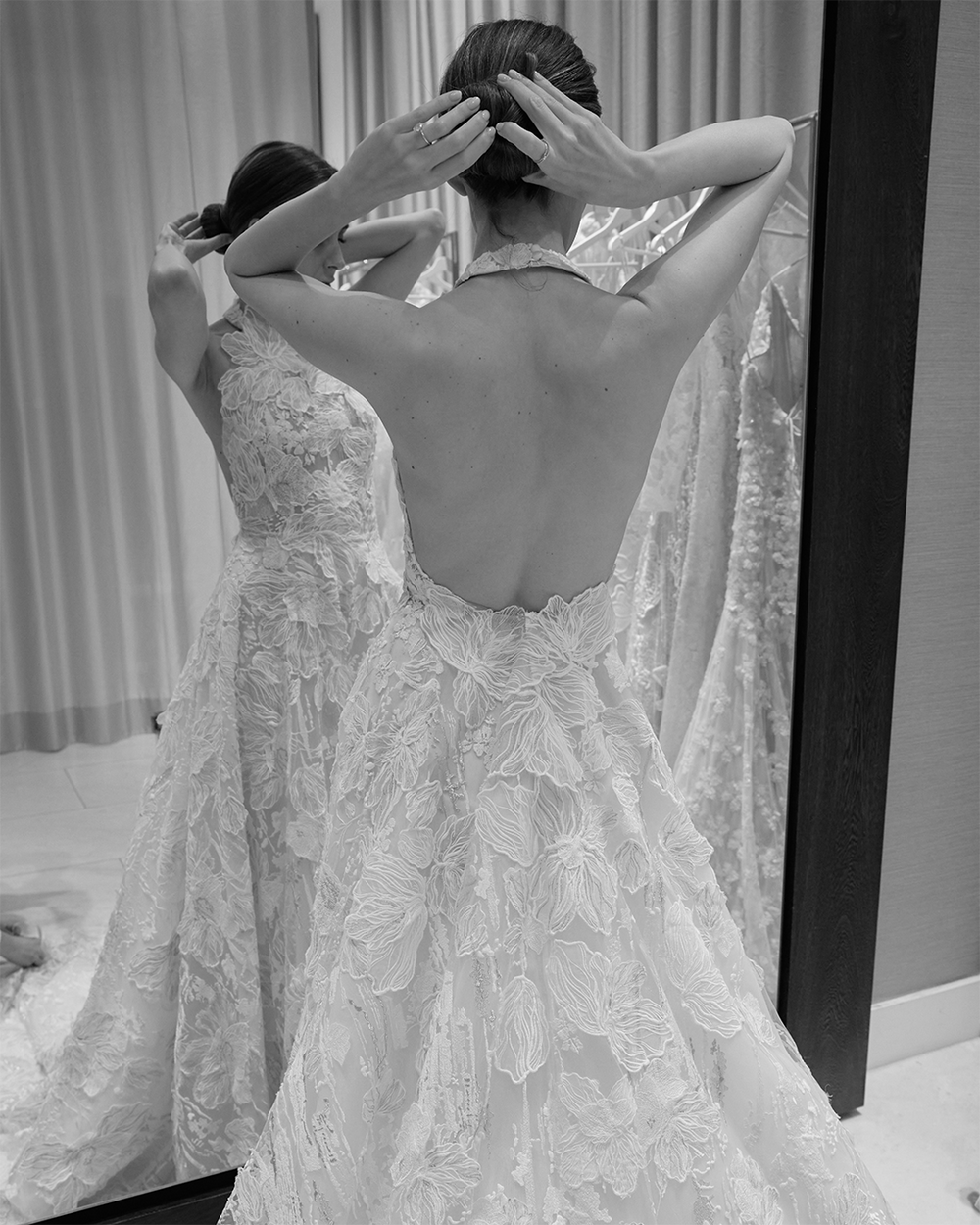 Una mujer con un vestido de novia se arregla el pelo delante de un espejo. El vestido de encaje tiene un estampado floral y la espalda abierta. Está de pie en un probador, rodeada de cortinas y otros vestidos.