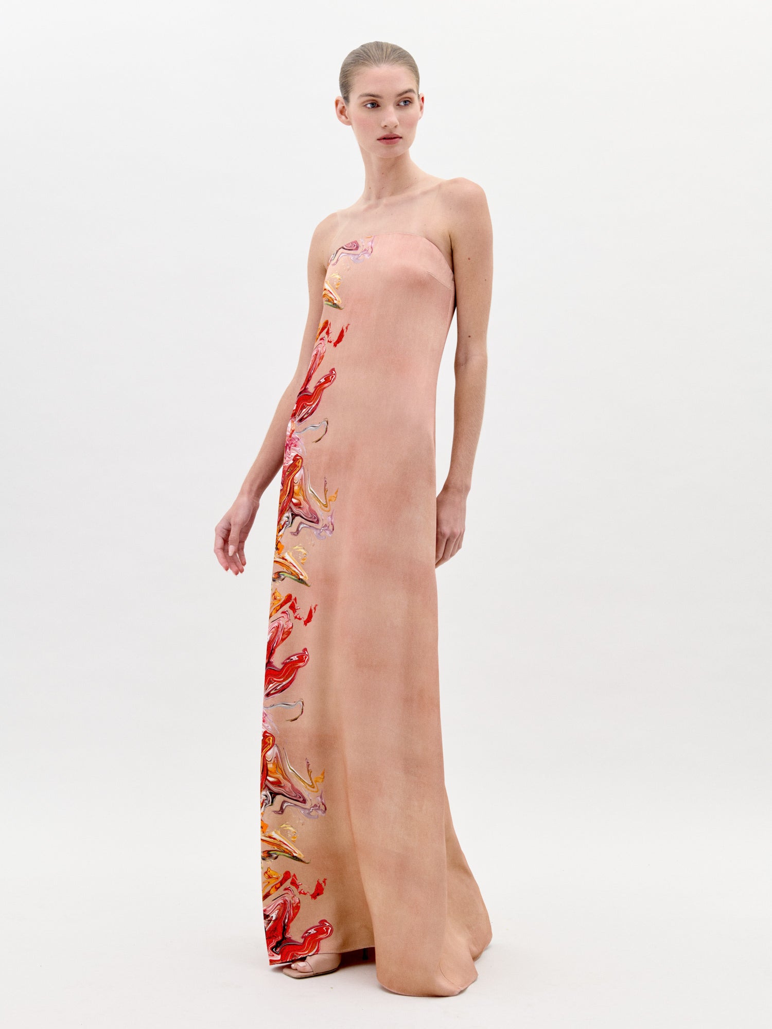 Una mujer se encuentra de pie sobre un fondo claro y liso con el Vestido Adalgisa, un vestido largo rosa pálido sin tirantes y largo hasta el suelo, con coloridos estampados abstractos en un lateral. Lleva el cabello recogido y la mirada ligeramente a la izquierda.