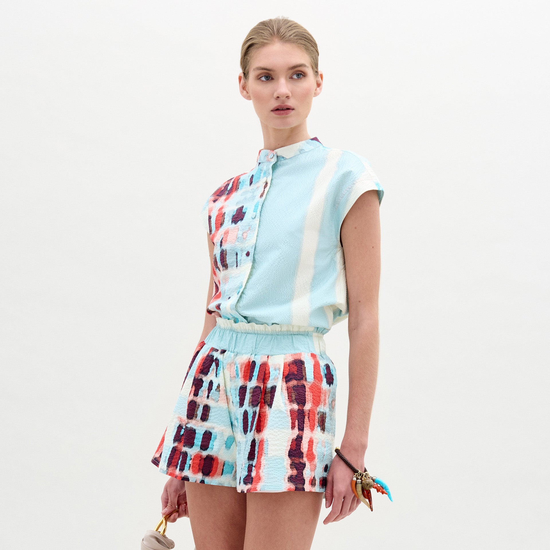 Una mujer posa sobre un fondo liso con la Blusa Adele, un top azul claro sin cuello, combinado con shorts geométricos a juego en rojo, azul y blanco. Lleva un pequeño bolso de mano y el cabello recogido. Hecho en Colombia con la mejor tela italiana para un estilo elegante.
