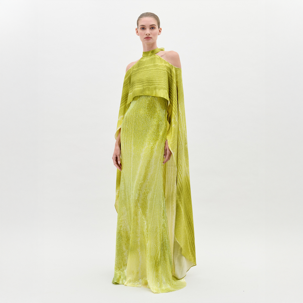 Una mujer posa relajada frente a un fondo blanco luciendo el Vestido Adelia, un maxi de lentejuelas amarillo-verde con cintura entallada, capa superpuesta y diseño de hombros descubiertos que realza su elegancia.