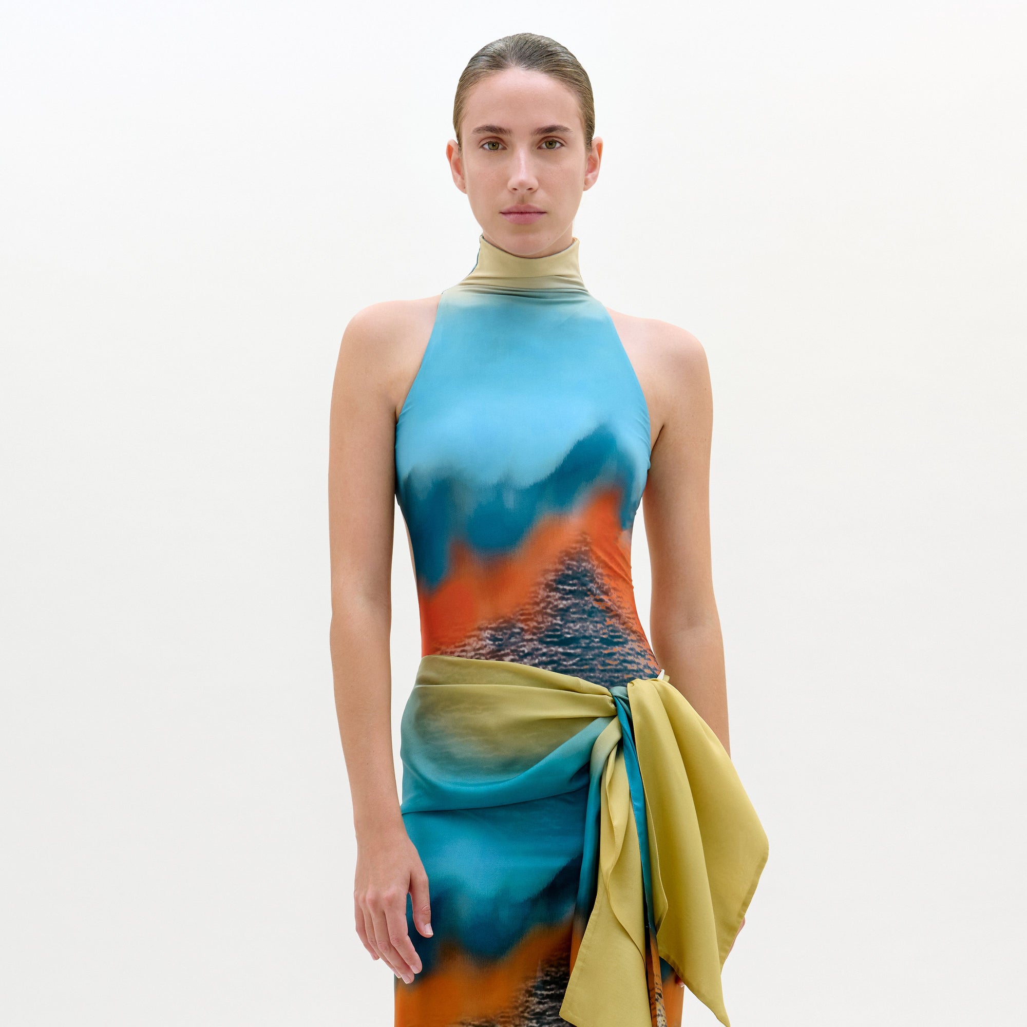 Una persona de pie, de frente, lleva el body Adina, una prenda sin mangas, de cuello alto, con un estampado abstracto azul, naranja y amarillo y un fajín amarillo a juego en la cintura. El fondo es liso y de color claro.