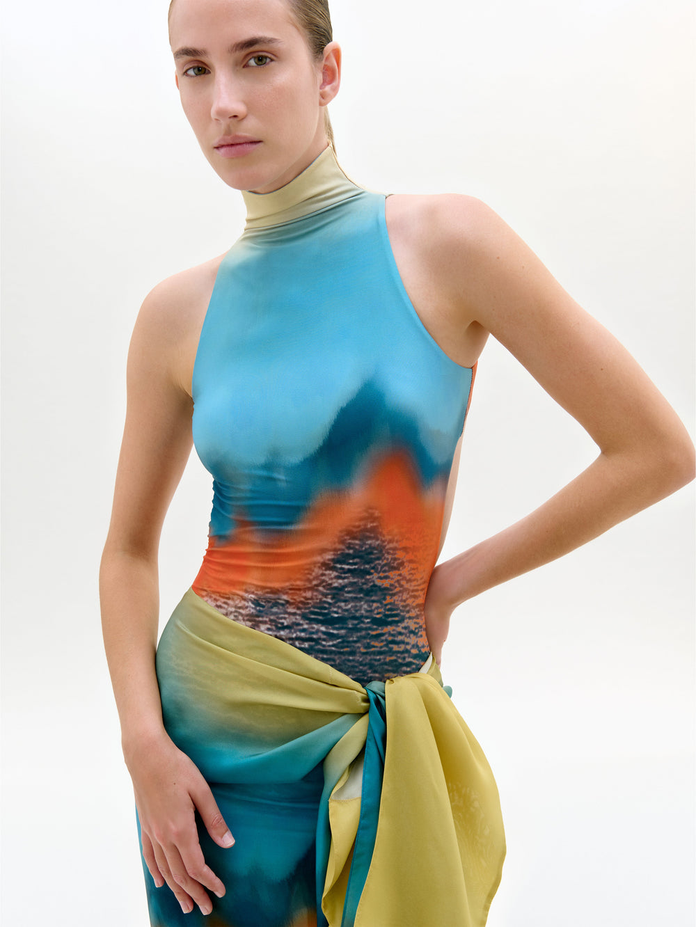 Una persona con el body Adina, con cuello alto, diseño sin mangas con estampado azul, naranja y agua en la parte superior y falda con detalle envolvente amarillo-verde, de pie sobre un fondo claro liso.