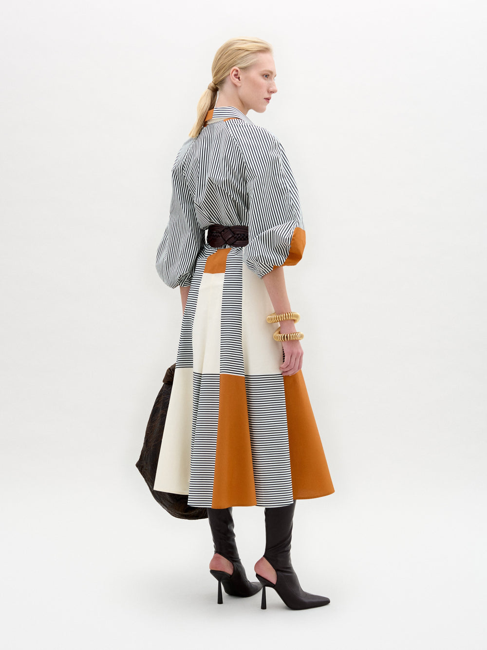 Una persona rubia con el pelo recogido lleva el vestido Agostina, una prenda midi de color con paneles marrones, blancos y naranjas, combinada con botas marrones de tacón alto y pulseras gruesas, sobre un fondo liso.