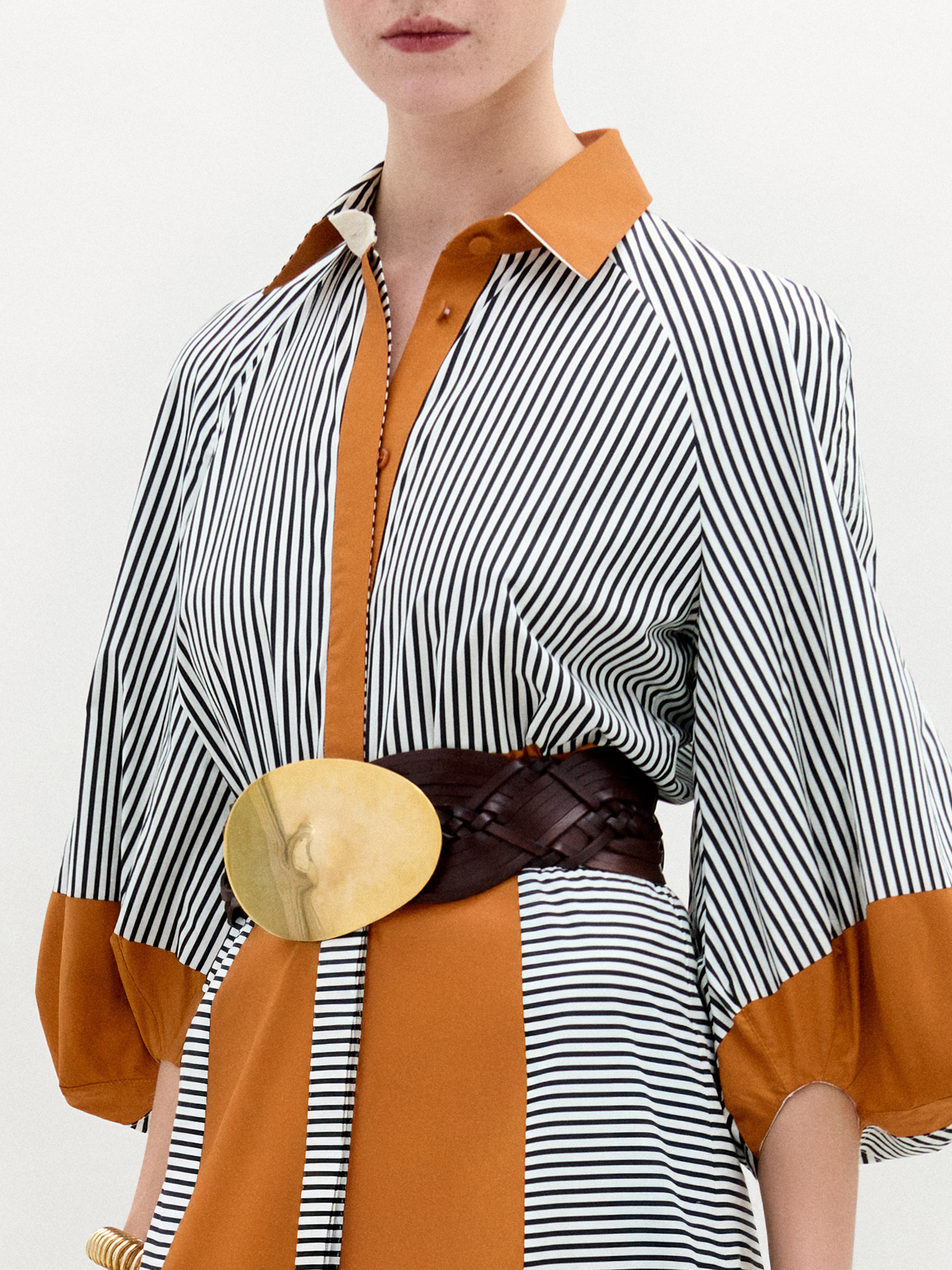 Una persona lleva el vestido Agostina, un midi de cuello con rayas verticales blancas y negras, paneles y puños de color naranja sólido, además de un cinturón marrón ancho con una gran hebilla dorada. Confeccionado en Colombia con tejido italiano. El rostro está parcialmente fuera del encuadre.