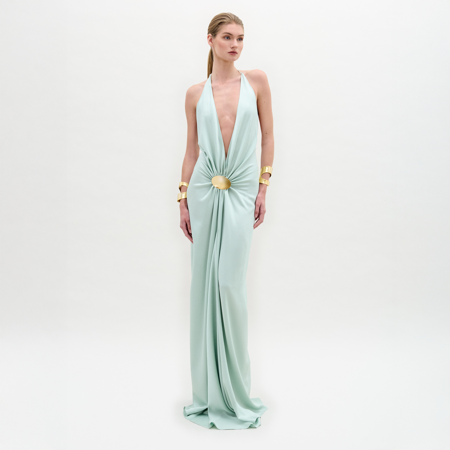 Sobre un fondo blanco liso, una persona luce el vestido Aiko, un maxi azul claro de silueta entallada, profundo escote en V, cintura fruncida con detalle circular dorado y combinado con brazaletes dorados en ambas muñecas.