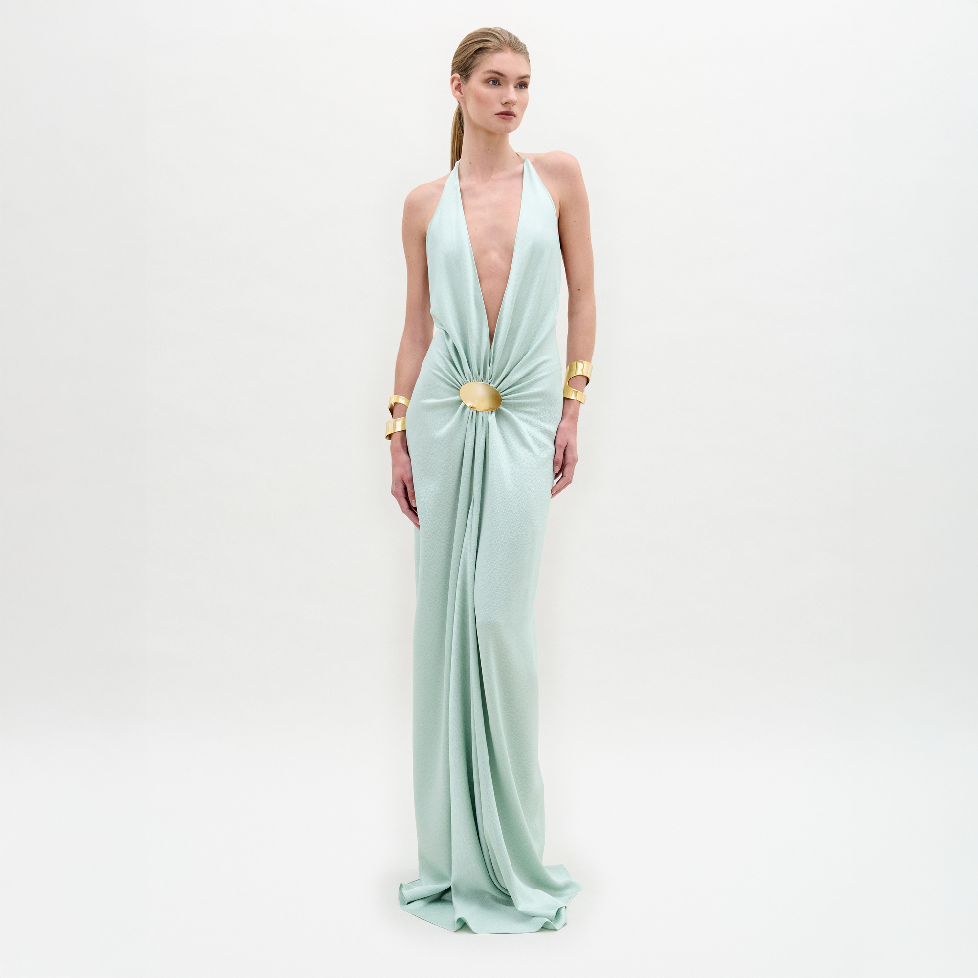 Sobre un fondo blanco liso, una persona luce el vestido Aiko, un maxi azul claro de silueta entallada, profundo escote en V, cintura fruncida con detalle circular dorado y combinado con brazaletes dorados en ambas muñecas.