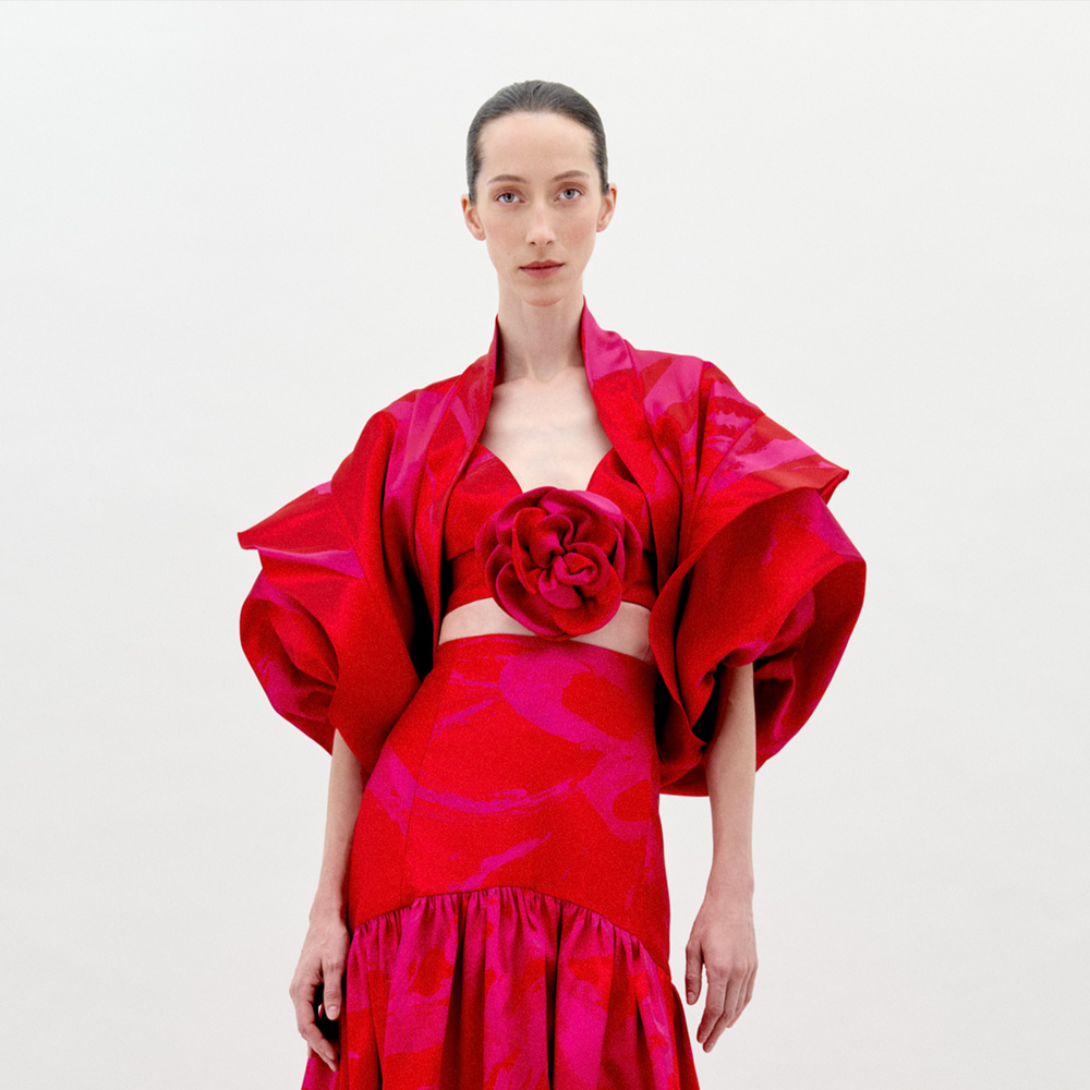 Una mujer viste el Shrug Aleen, una llamativa pieza de tafetán rojo y rosa con mangas voluminosas, un gran detalle de estampado floral en el pecho y una falda de cintura alta, de pie sobre un fondo blanco liso.