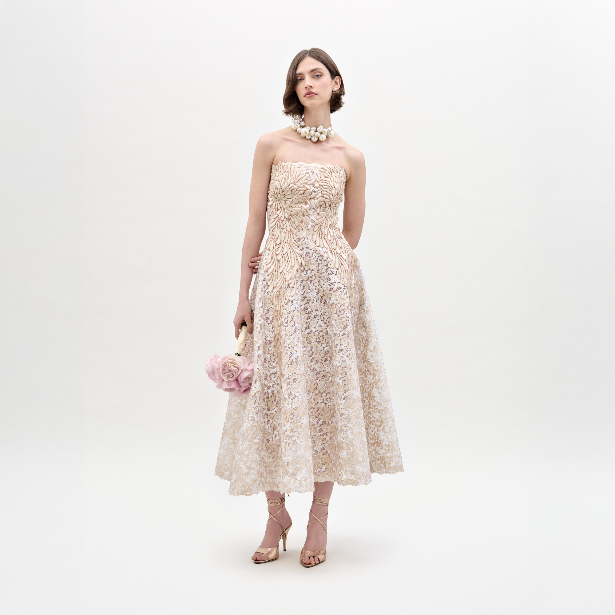 Una mujer con un vestido Alejandra beige de Atelier Tela Italiana se encuentra sobre un fondo blanco, adornada con joyas de perlas, sandalias de tacón y sosteniendo flores rosas.