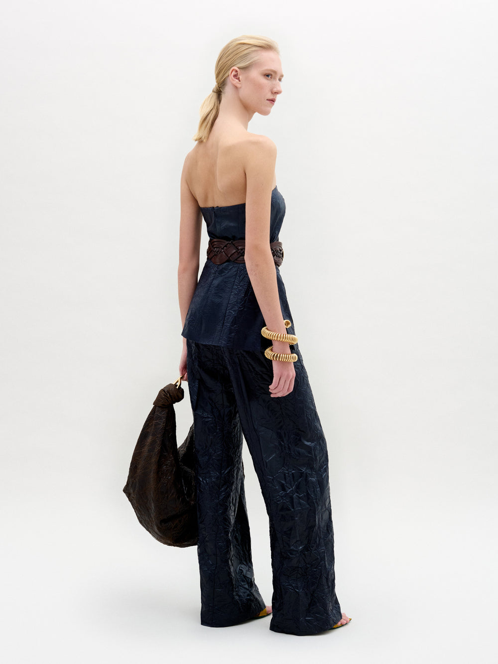 Sobre un fondo claro y liso, una mujer lleva el pantalón Emine, azul oscuro, de talle alto y pierna ancha, combinado con un top de tirantes a juego. Lleva un bolso grande oscuro, pulseras doradas gruesas y el pelo recogido.