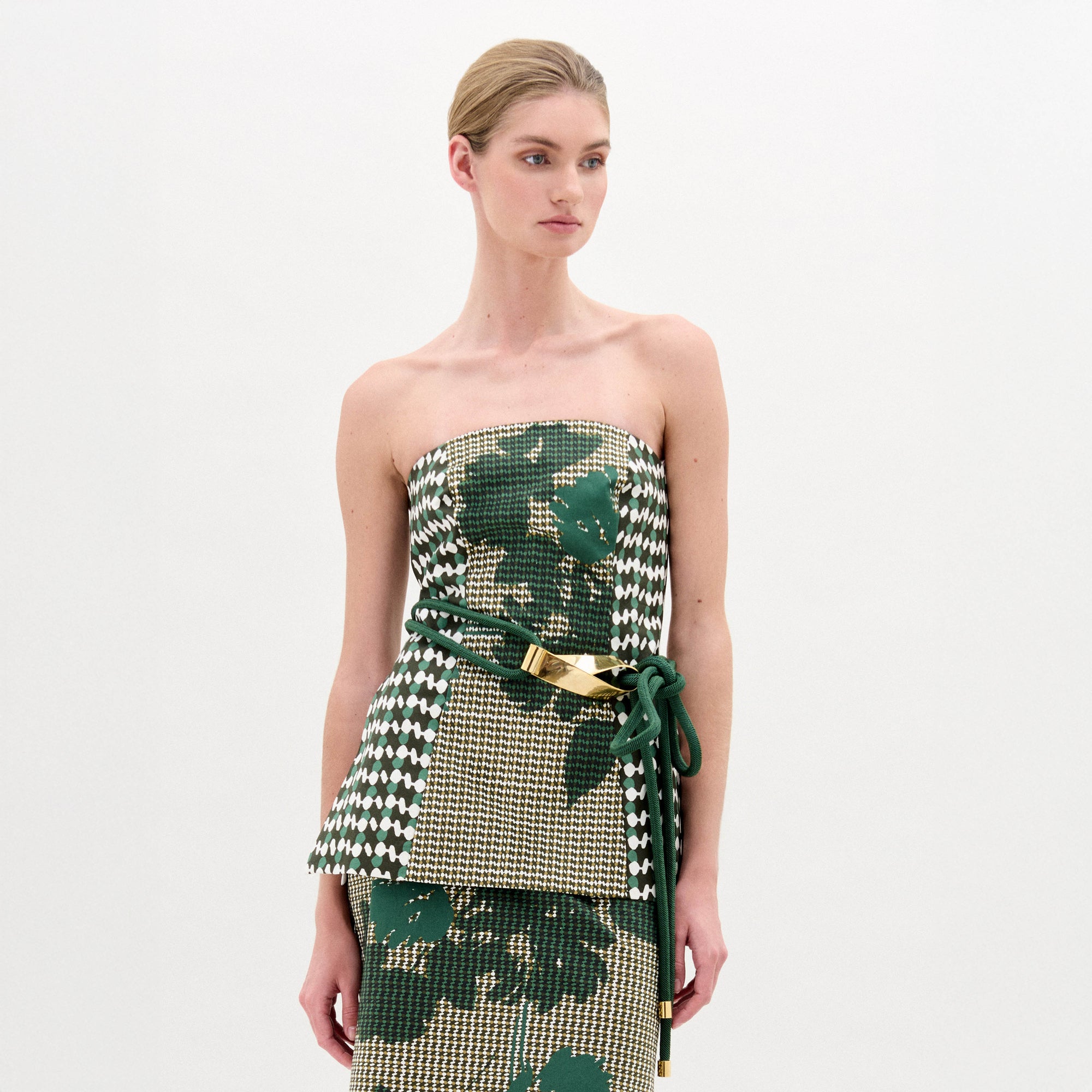 Una mujer con el pelo rubio recogido en un moño bajo lleva el top Astor y la falda a juego con estampados de hojas y pata de gallo en verde y beige, rematados con un cinturón de cuerda verde con detalles metálicos. Sobre fondo blanco liso. Hecho en Colombia.