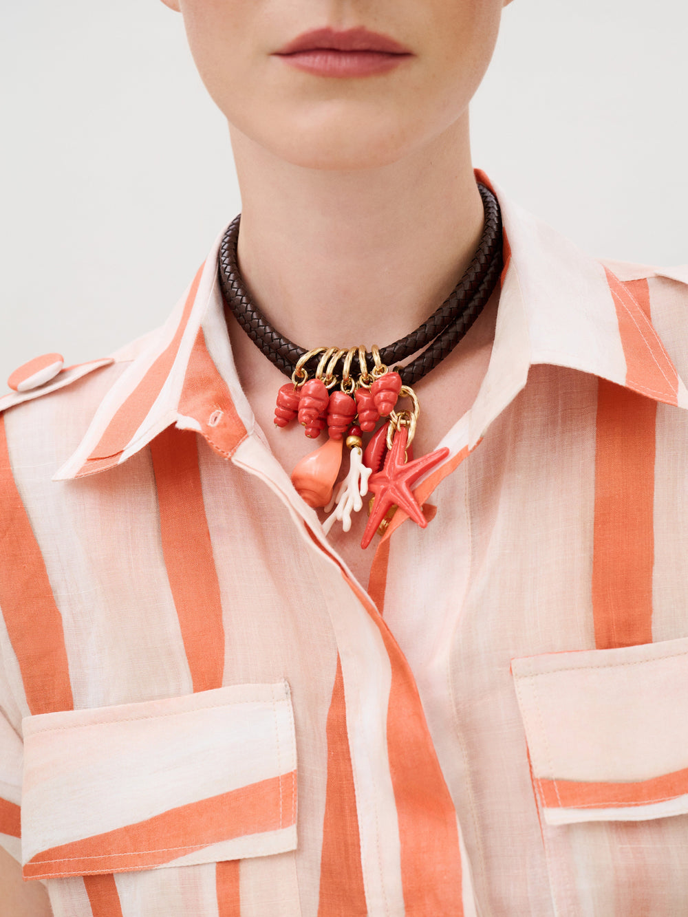 A person wearing an Aaron Romper in Italian fabric and a brown braided leather necklace featuring red and pink charms shaped like coral, shells, and a starfish.