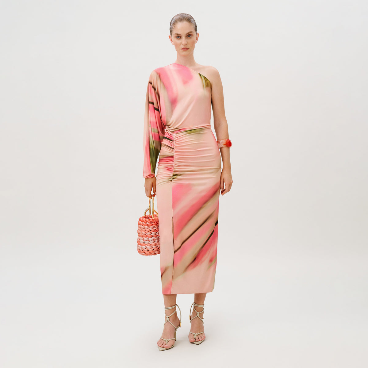 A model wears the Albany Dress, an asymmetrical, one-shoulder Lycra midi dress featuring a pink and green abstract print with ruched detailing, accessorized with strappy heels and a woven handbag.