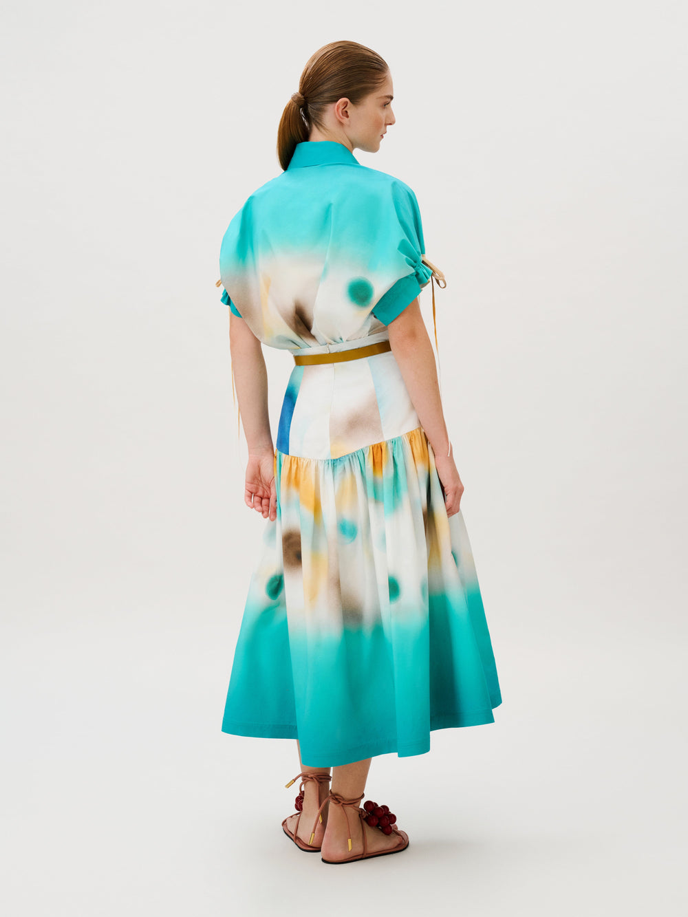A person wearing the Aliana Skirt, a 100% cotton midi-length dress in a teal and yellow tie-dye pattern, featuring puffed sleeves with tie details and a mustard-colored belt.