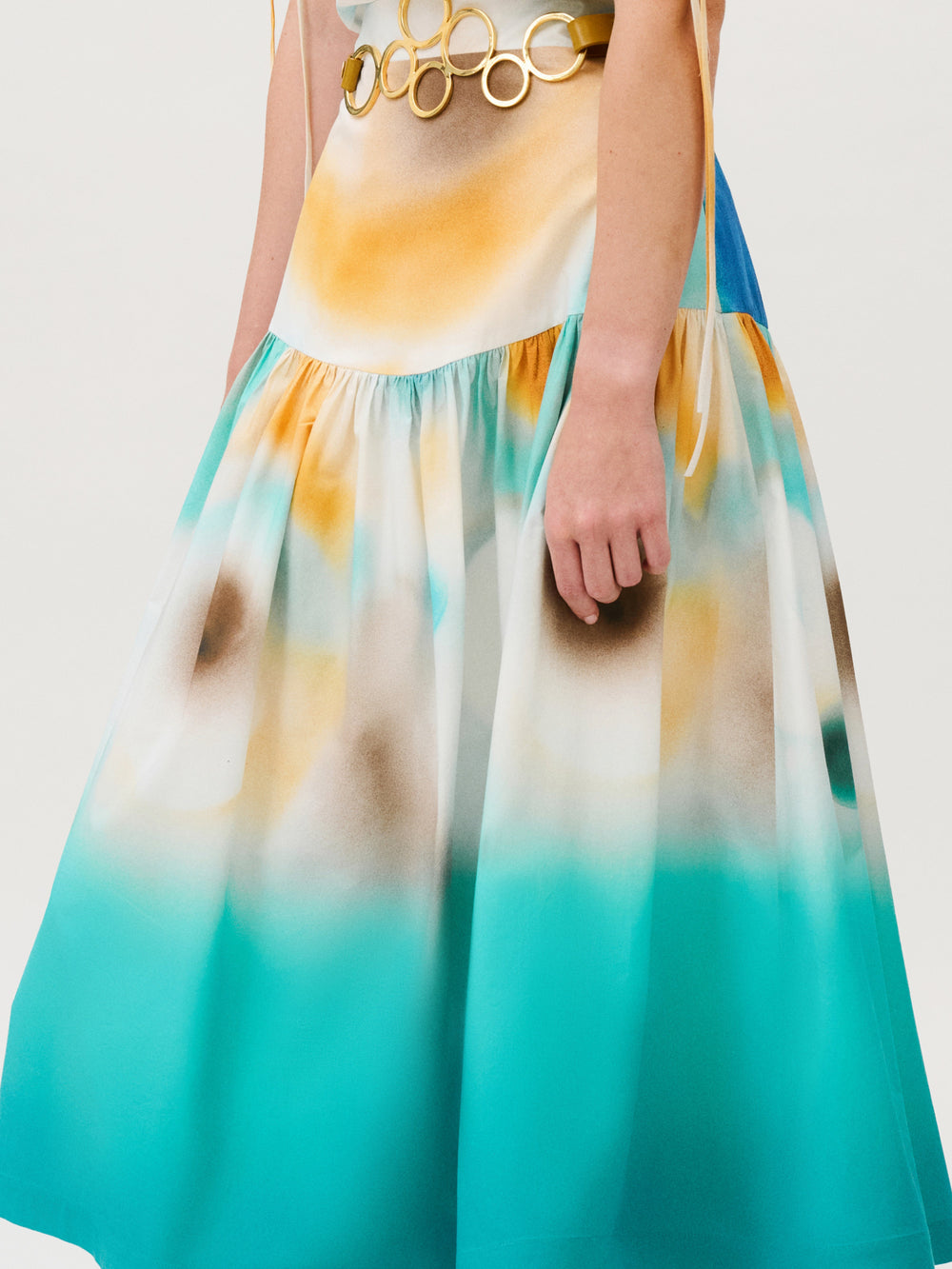 The Aliana Skirt is a midi skirt made from Italian fabric with a tie-waist featuring large gold rings and a blue, yellow, and white airbrushed pattern.