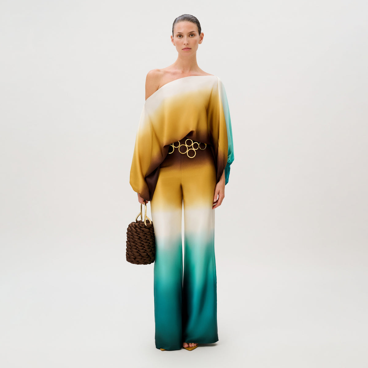 A person wearing an Andie Pant silk jumpsuit featuring an off-the-shoulder, ombre-patterned top in shades of gold, brown, and teal, cinched with a gold-ringed belt and holding a dark brown woven handbag.