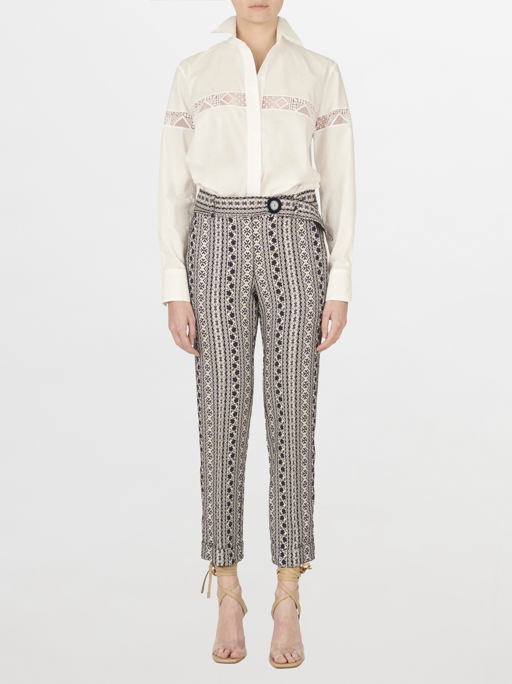 Una persona está de pie mirando hacia adelante con la Blusa Andrea White, que presenta un bordado intrincado, combinada con pantalones estampados de cintura alta y sandalias de tacón con cordones de color beige sobre un fondo blanco liso.