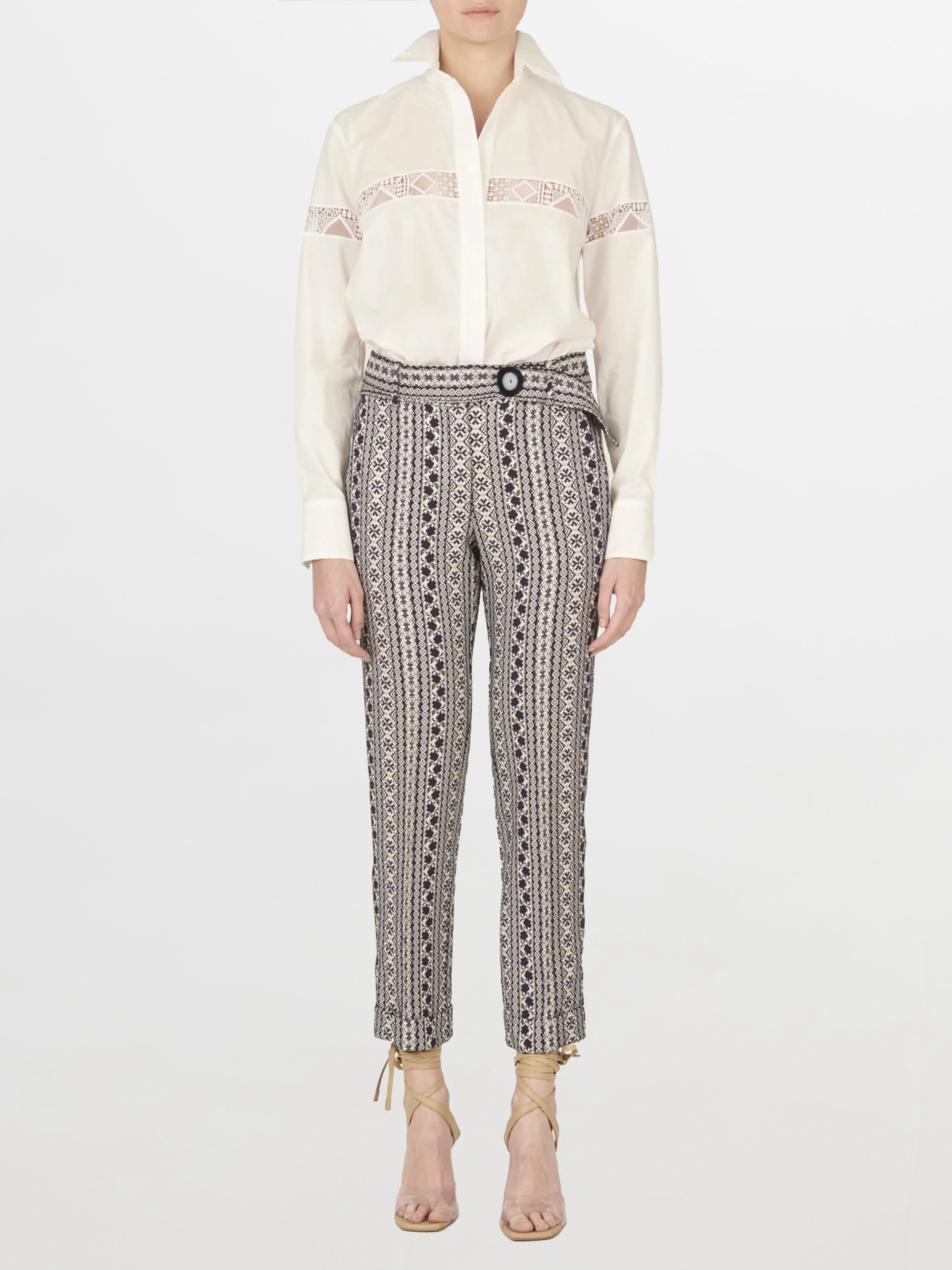 Una persona está de pie mirando hacia adelante con la Blusa Andrea White, que presenta un bordado intrincado, combinada con pantalones estampados de cintura alta y sandalias de tacón con cordones de color beige sobre un fondo blanco liso.