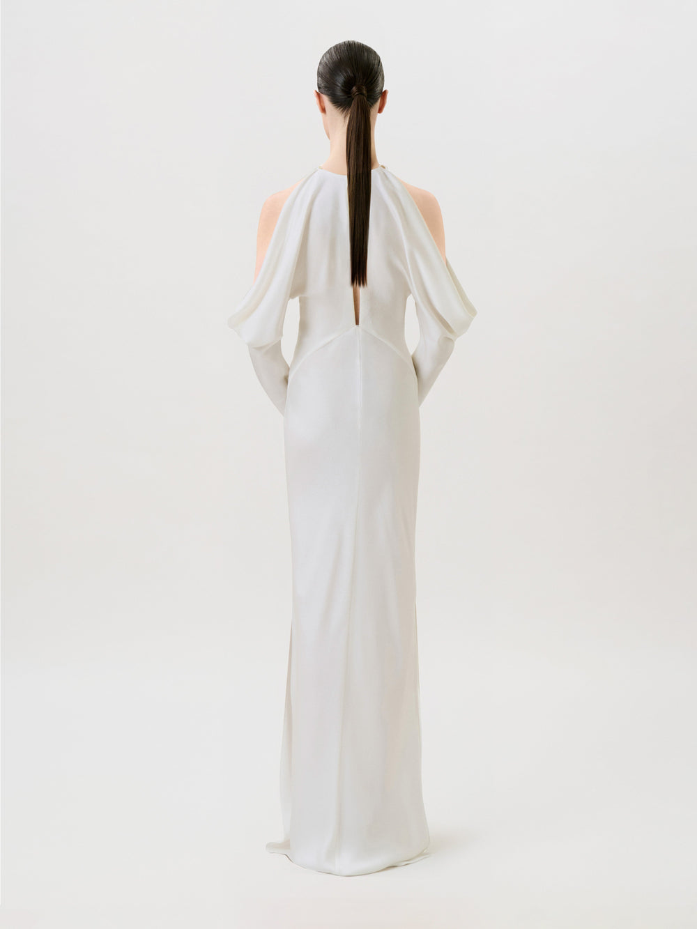A woman with straight dark hair in a ponytail models the Antonia Dress, crafted in hammered satin and designed with open shoulders. Posed with her back to the camera against a simple light background, she captures the essence of modern ready-to-wear sophistication.
