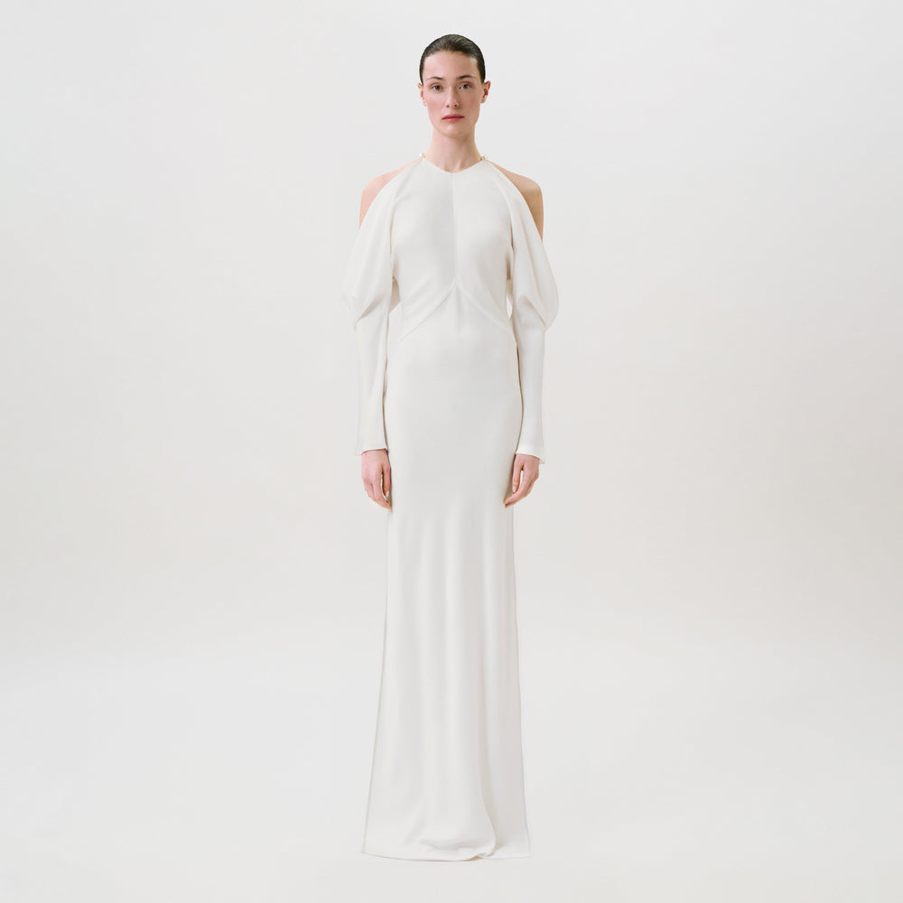 A person stands facing forward wearing the Antonia Dress—an elegant, long white gown featuring cut-out shoulders, long sleeves, and a fitted silhouette crafted from luxurious hammered satin—all set against a plain white background. This Ready To Wear piece exudes modern sophistication.