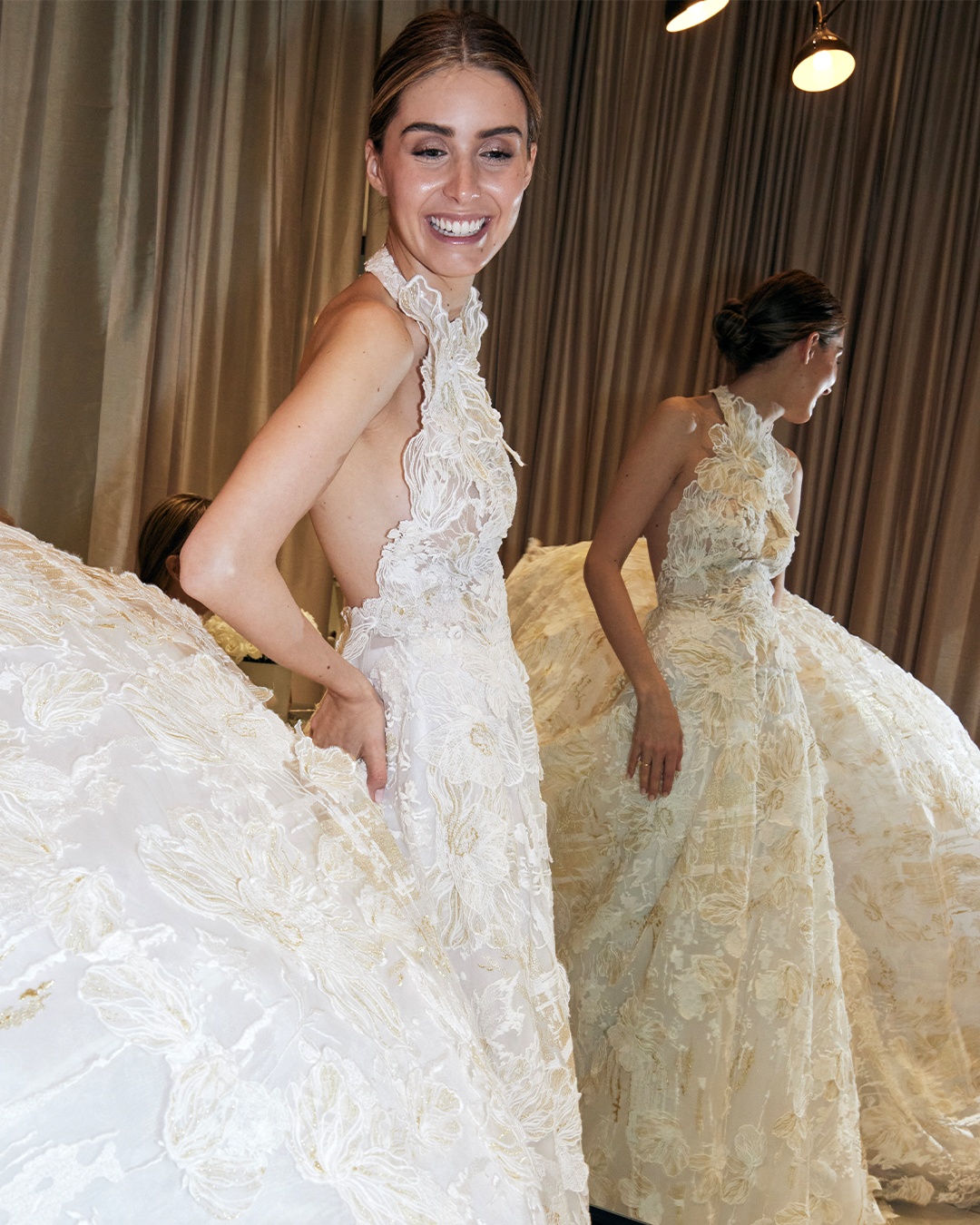 Una mujer con un elegante vestido de novia de encaje sin mangas está de pie sonriendo. El vestido presenta un intrincado bordado floral y un escote halter. En el fondo, se ve su reflejo en un espejo, lo que resalta la voluminosa cola del vestido.