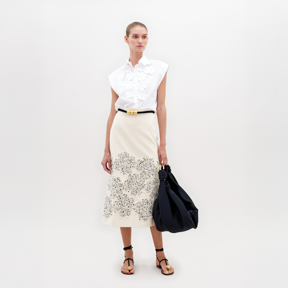 Una persona está parada sobre un fondo liso con una blusa blanca sin mangas con detalles de volantes y una Falda Atira beige con un diseño floral aplicado. Sostiene un bolso negro grande, usa sandalias negras y se complementa con un cinturón negro con una hebilla dorada.