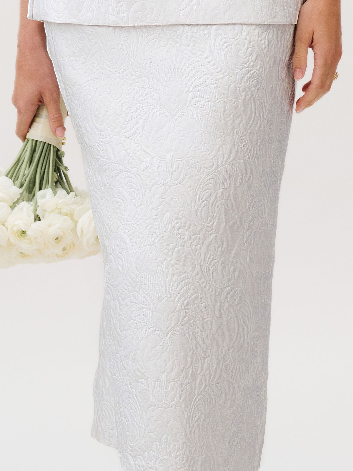 A person stands against a plain background wearing the Atira Skirt, an elegant, textured white piece crafted from luxurious Italian fabric, while holding a bouquet of white flowers in their left hand.