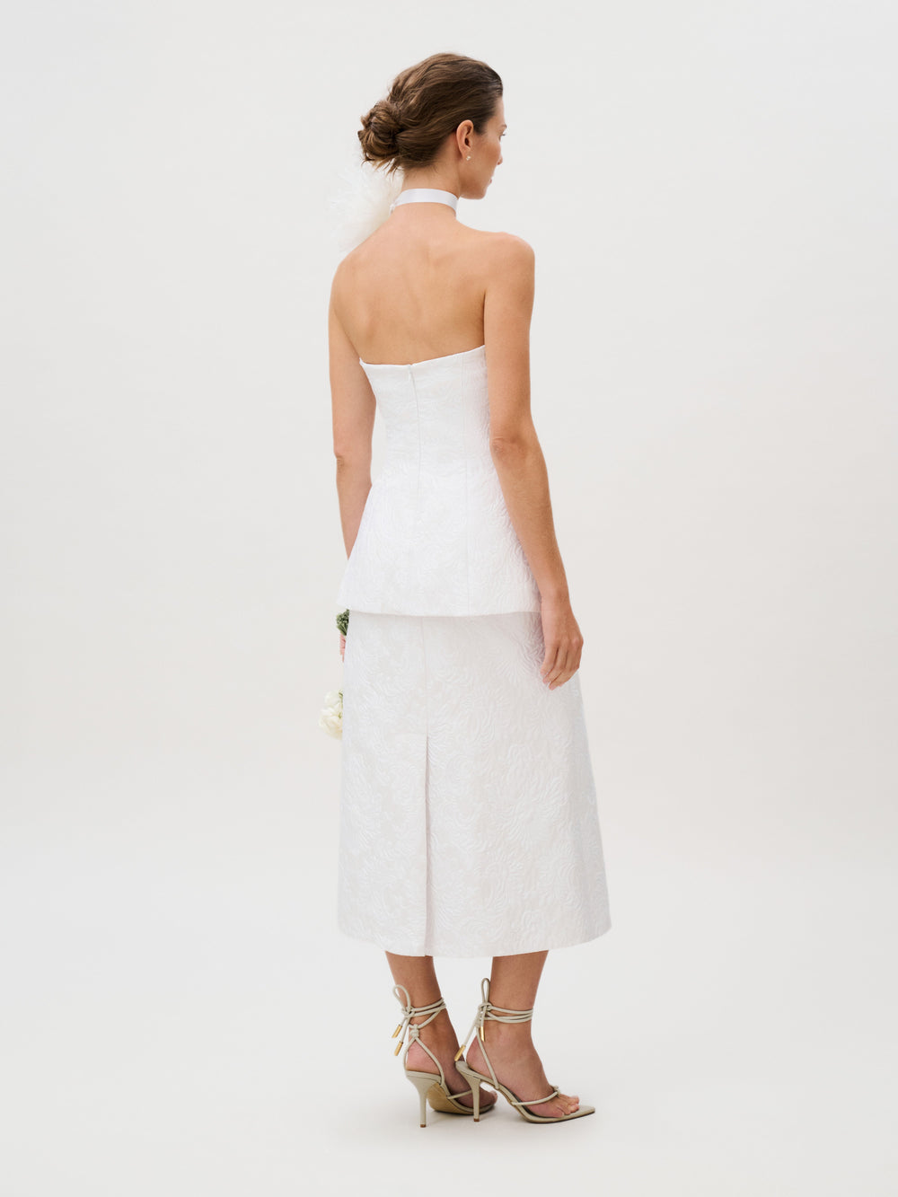 The woman wears the Atira Skirt, designed in a luxurious Italian white fabric with a strapless, halter neckline. She stands with her back to the camera, pairing the skirt with strappy heels and an elegant updo against a plain, light background.