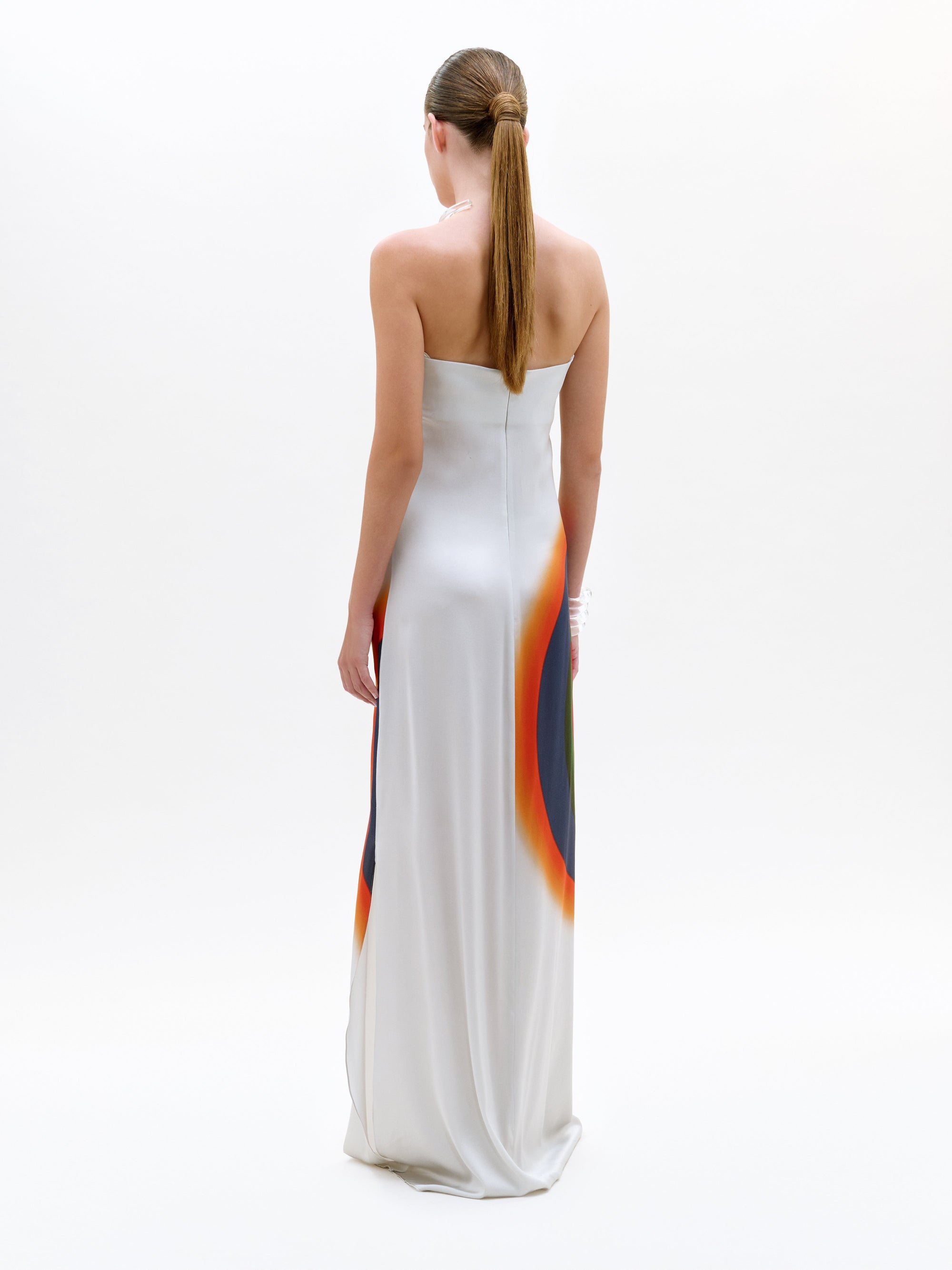 Una mujer con el pelo largo y liso recogido en una coleta baja, de pie y de espaldas, lleva el vestido Bahiana, un maxi de seda hasta el suelo, sin tirantes, con degradados circulares de colores a los lados. Fondo blanco liso. Fabricado en Portugal.